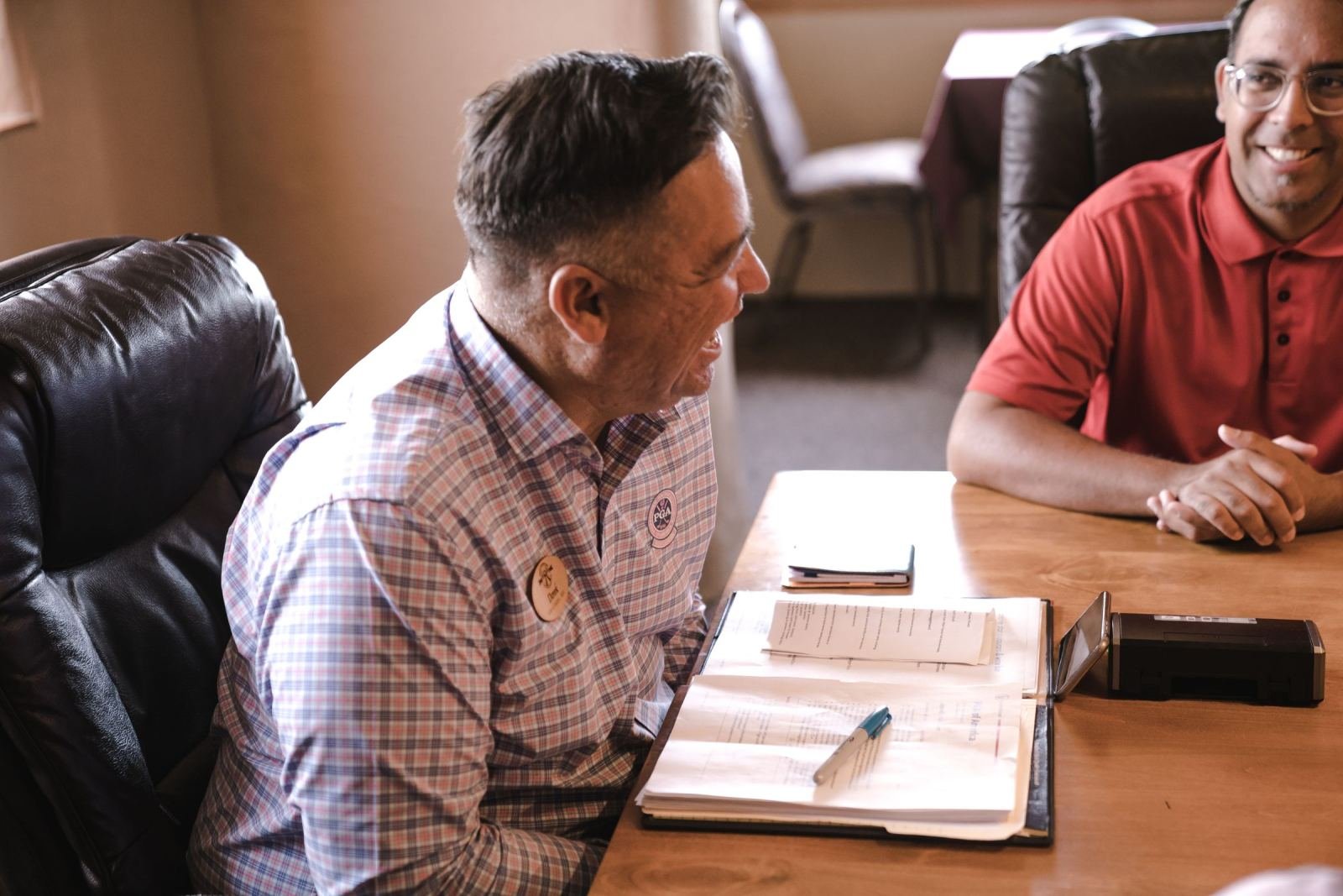 Two men discuss paperwork while seated at a desk in a well-lit office.