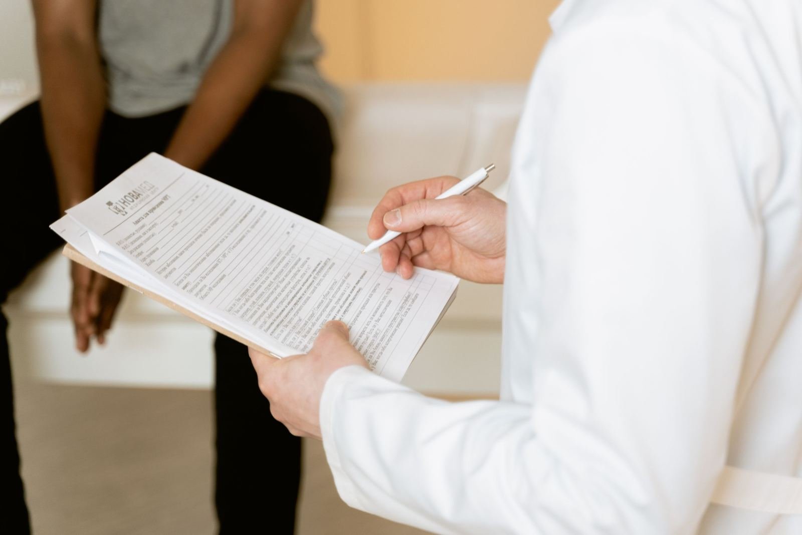 A patient sits behind a clinician in a white coat who is writing on a clipboard