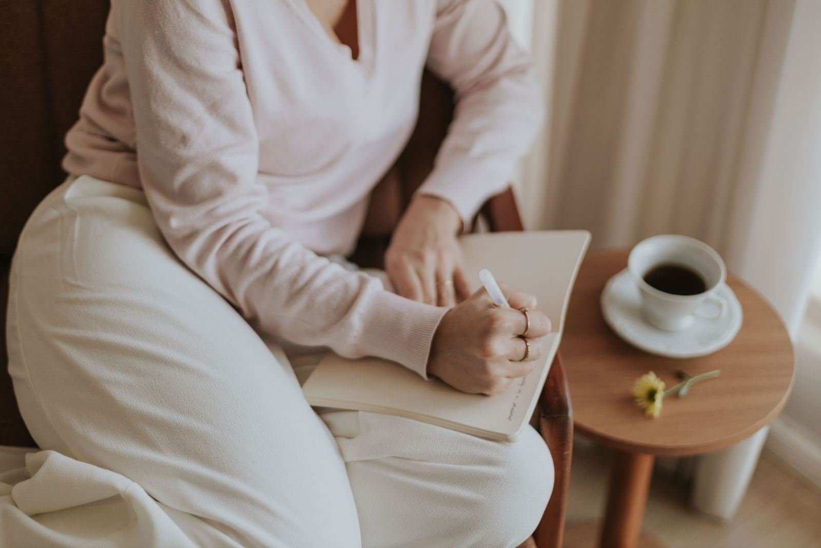 A person in a pink sweater writes in a journal next to a cup of coffee