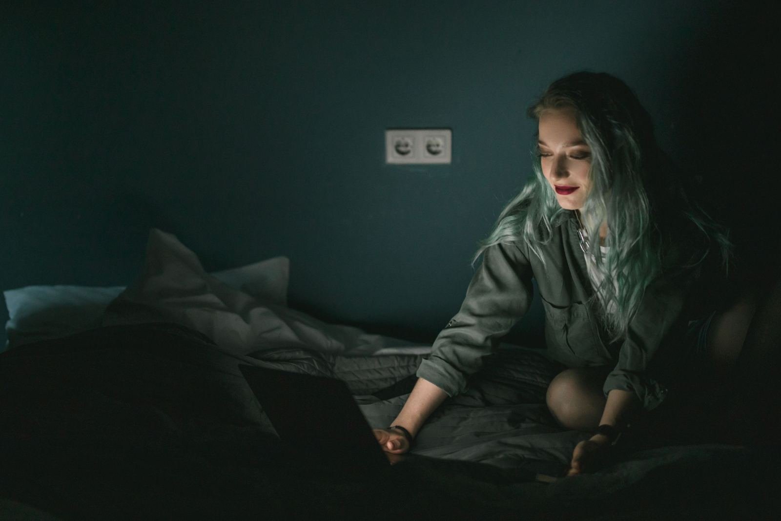 A woman with blue hair looks at a laptop screen in a dark room