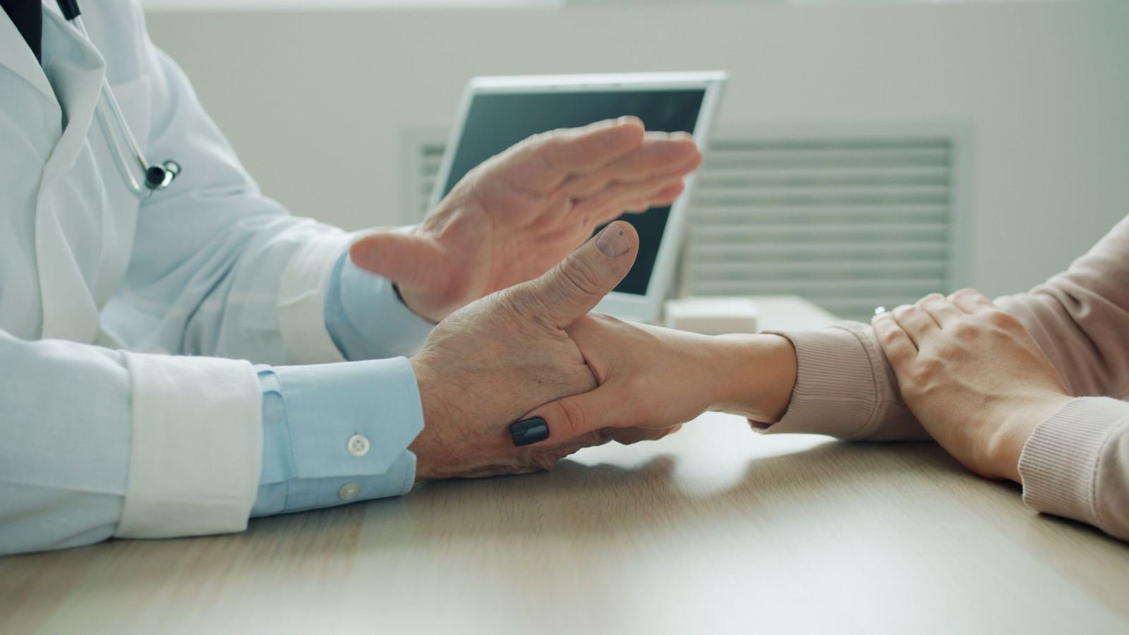 A clinician in a white coat holds a patient's hand across a desk