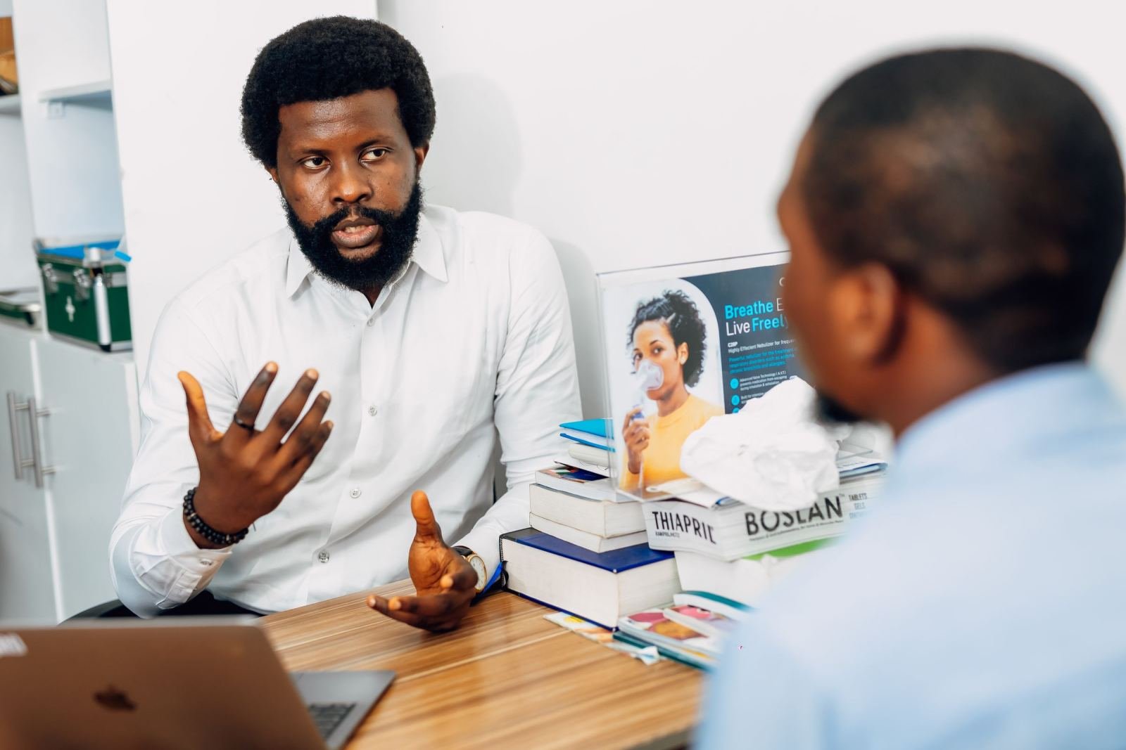 Two people talk across a desk. One wears a white shirt and makes a hand gesture.