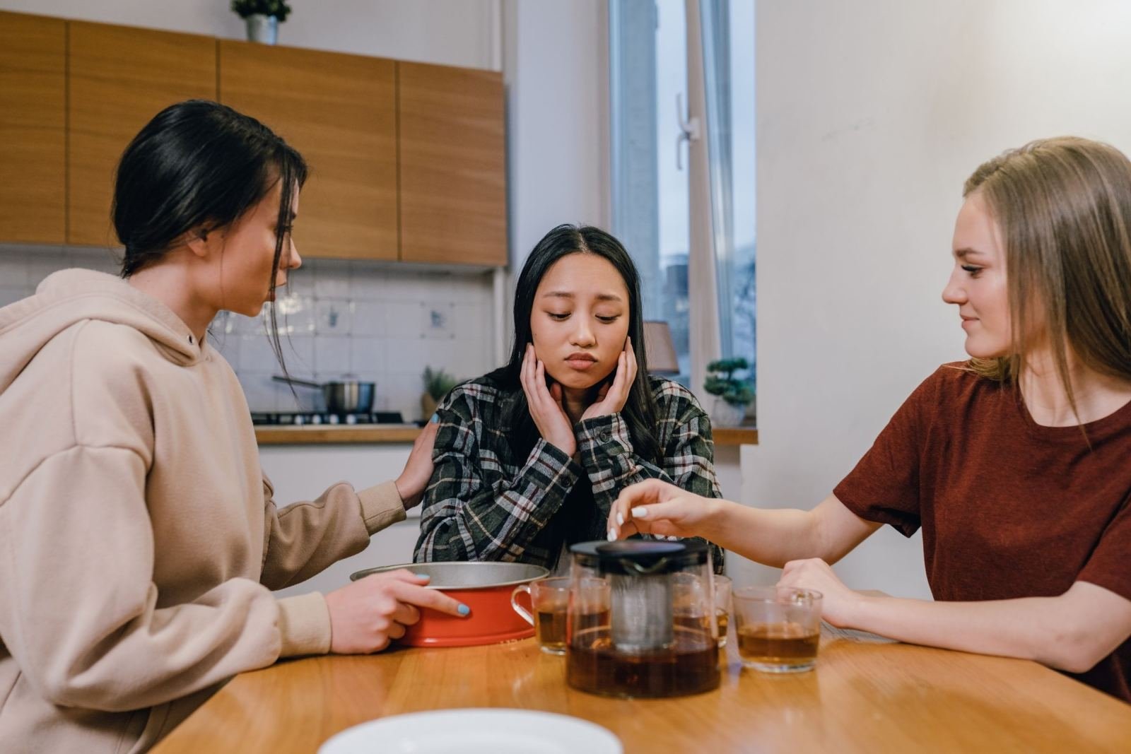 Three women sit at a kitchen table drinking tea. They are supporting a woman who looks sad.
