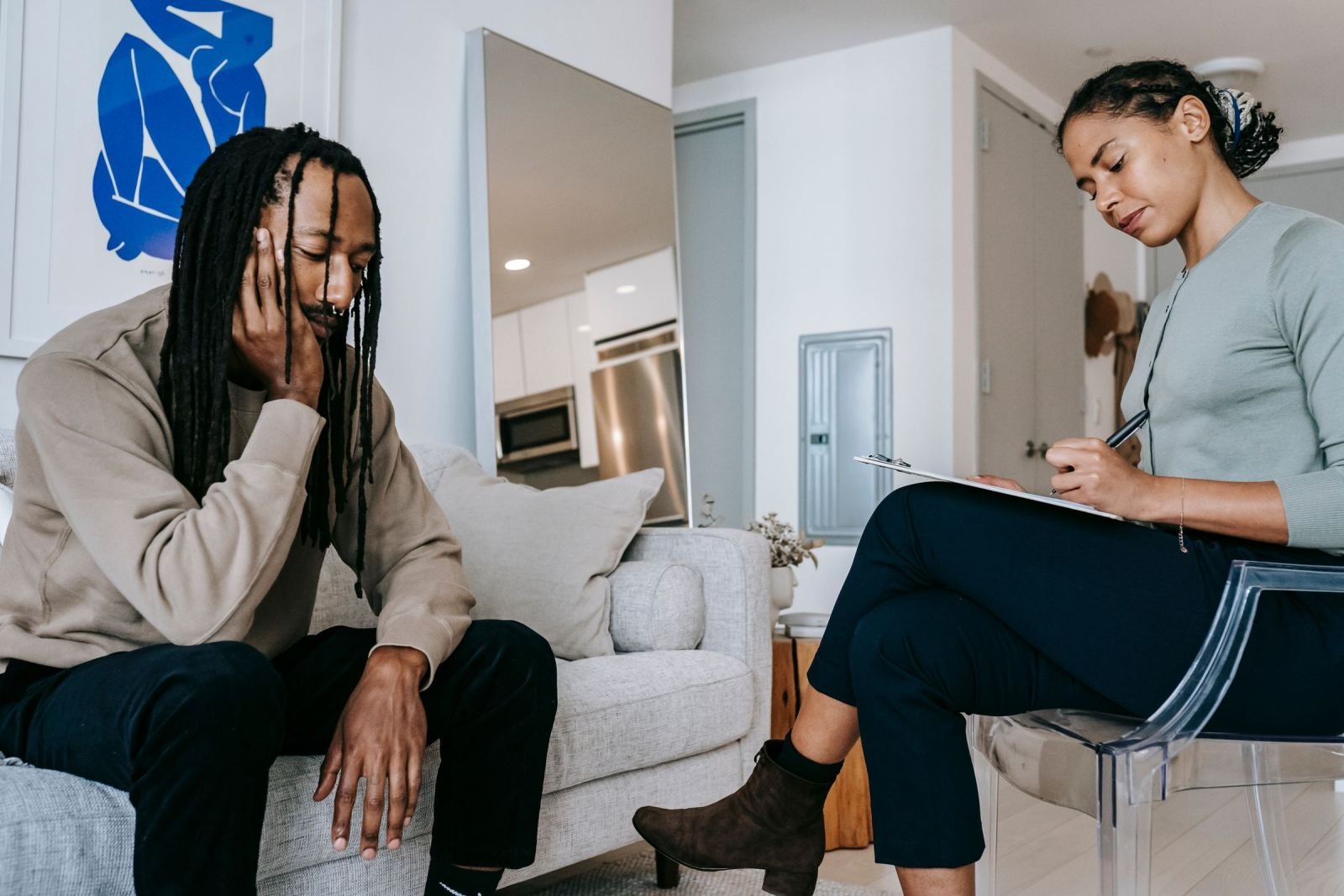 A woman with a clipboard takes notes while talking to a man on a sofa