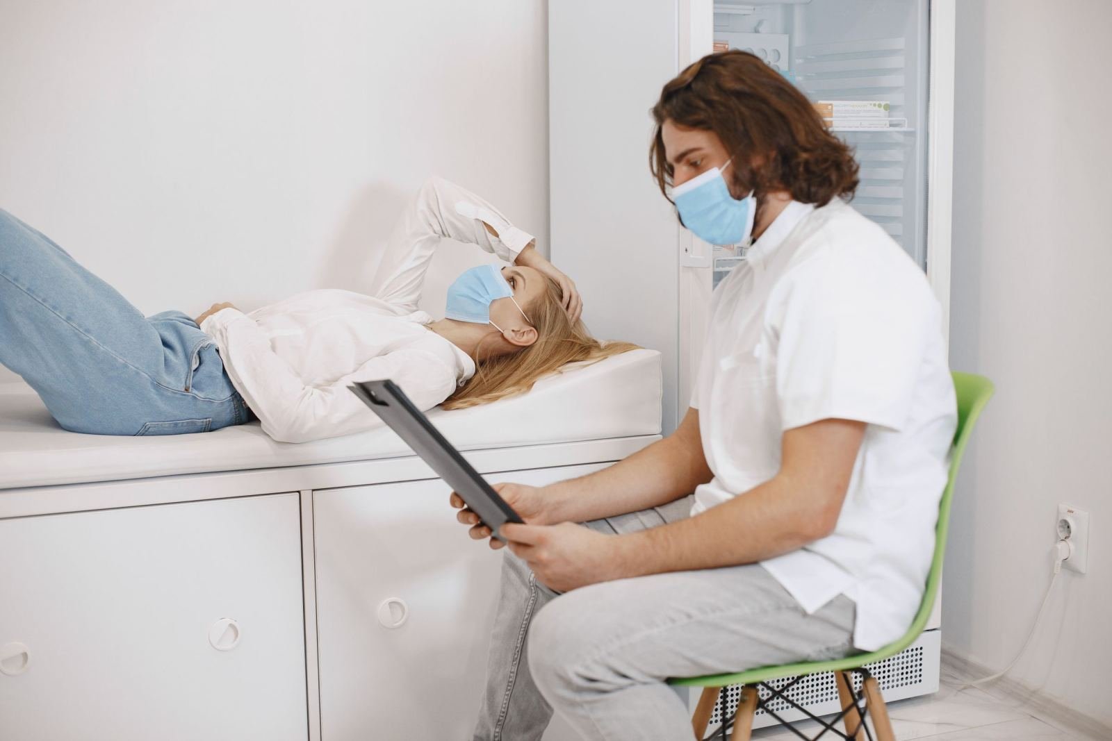 A masked clinician speaks with a patient on an exam table, holding a clipboard