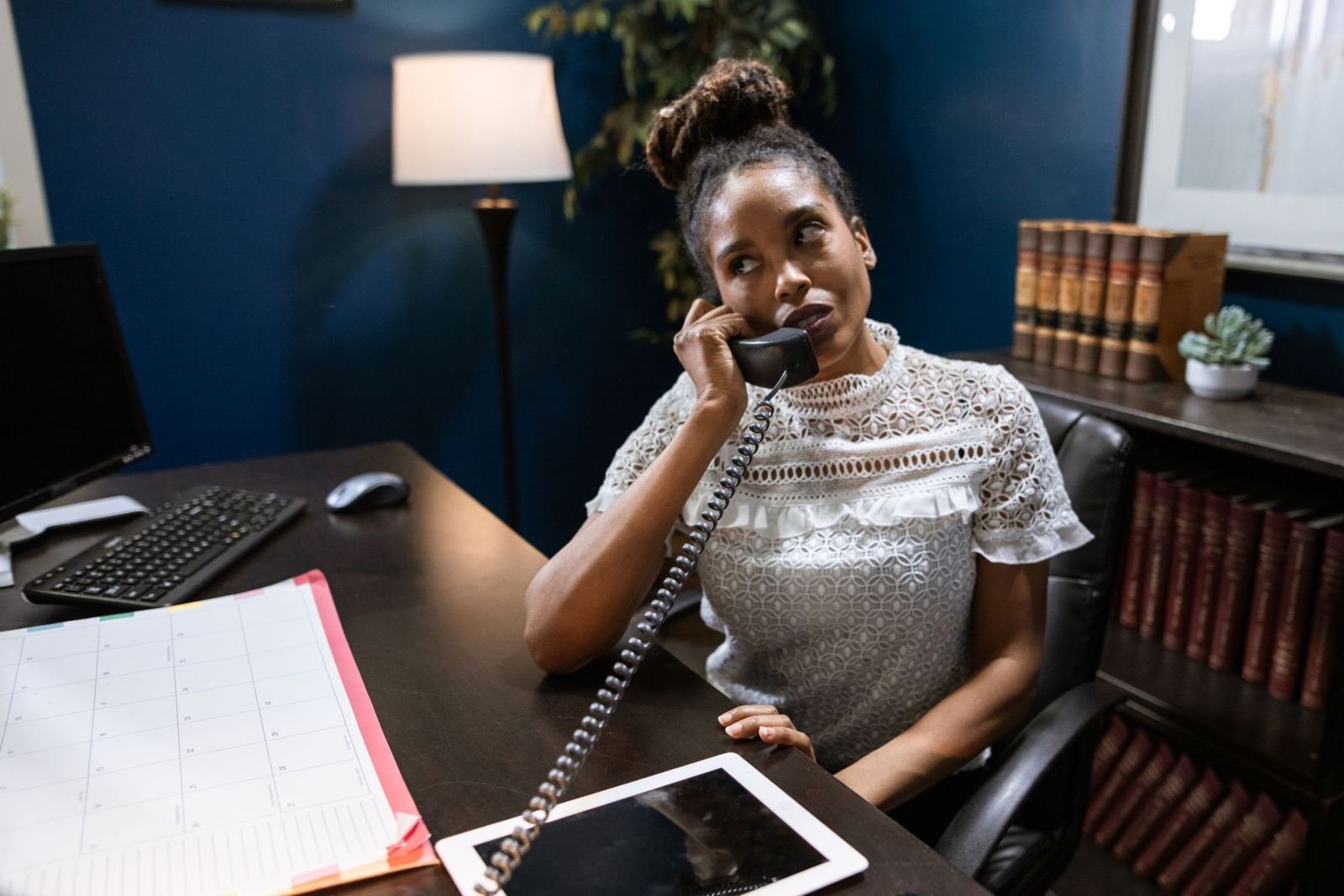 A woman in a white lace top speaks on a corded phone at her office desk