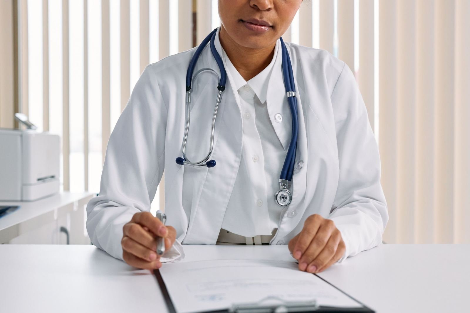 A clinician in a white coat and stethoscope looks at a clipboard on a desk