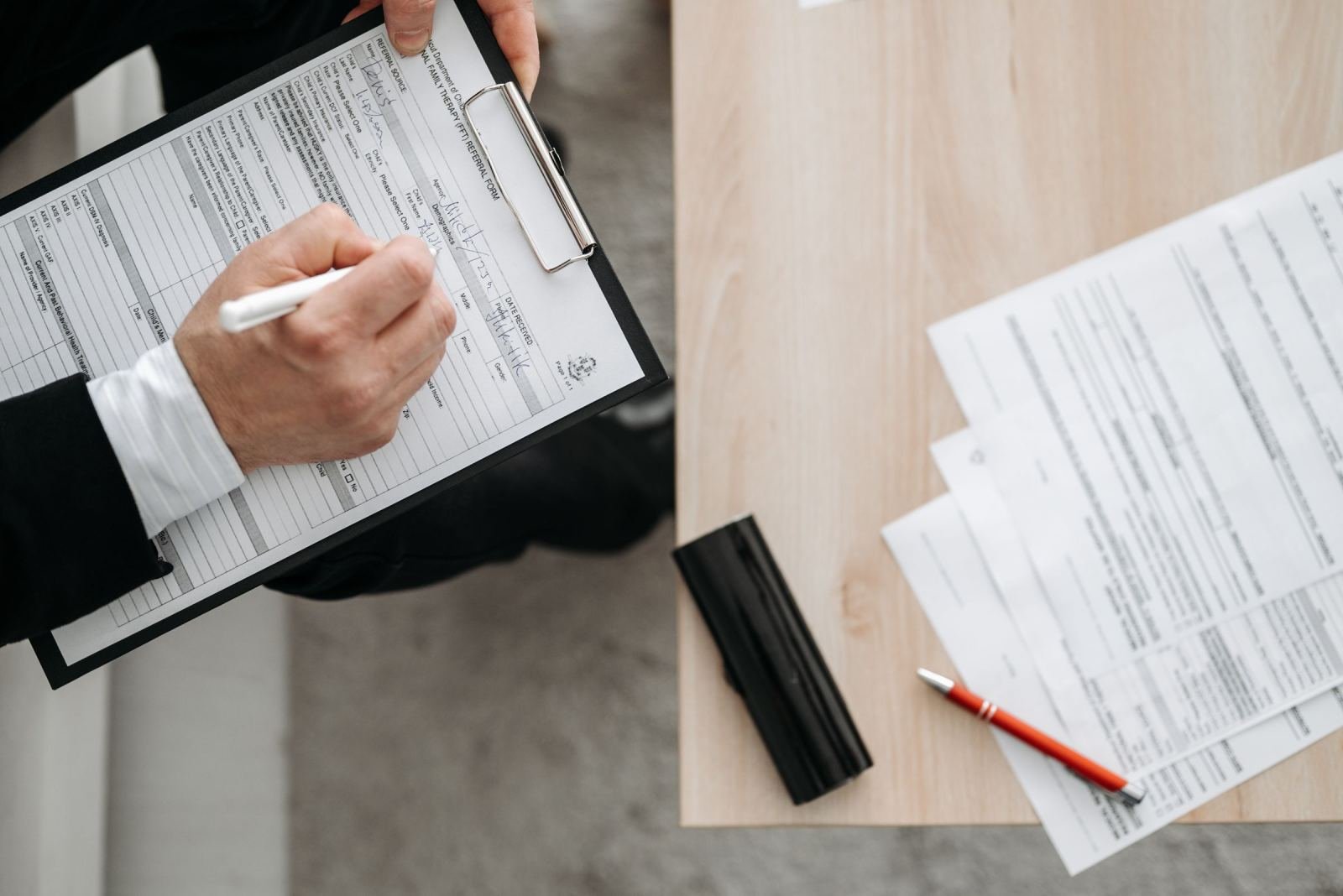 A person in a suit fills out a form on a clipboard at a wooden desk