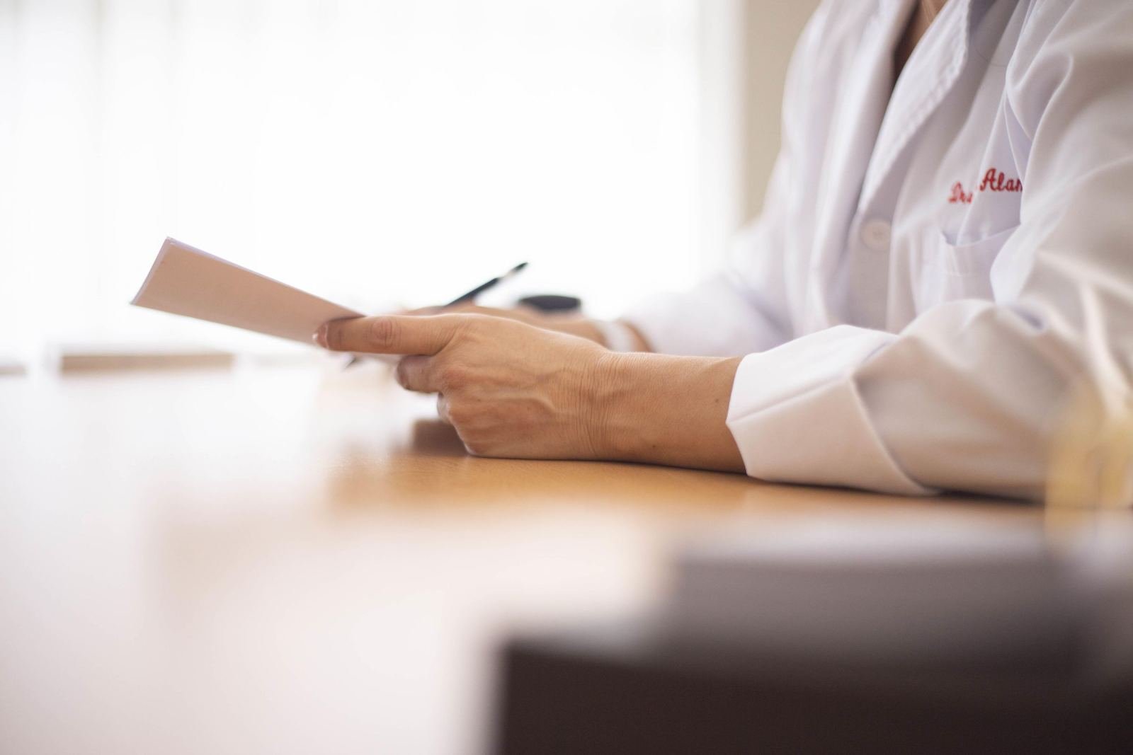 A person wearing a white lab coat stands at a wooden desk holding a small white card