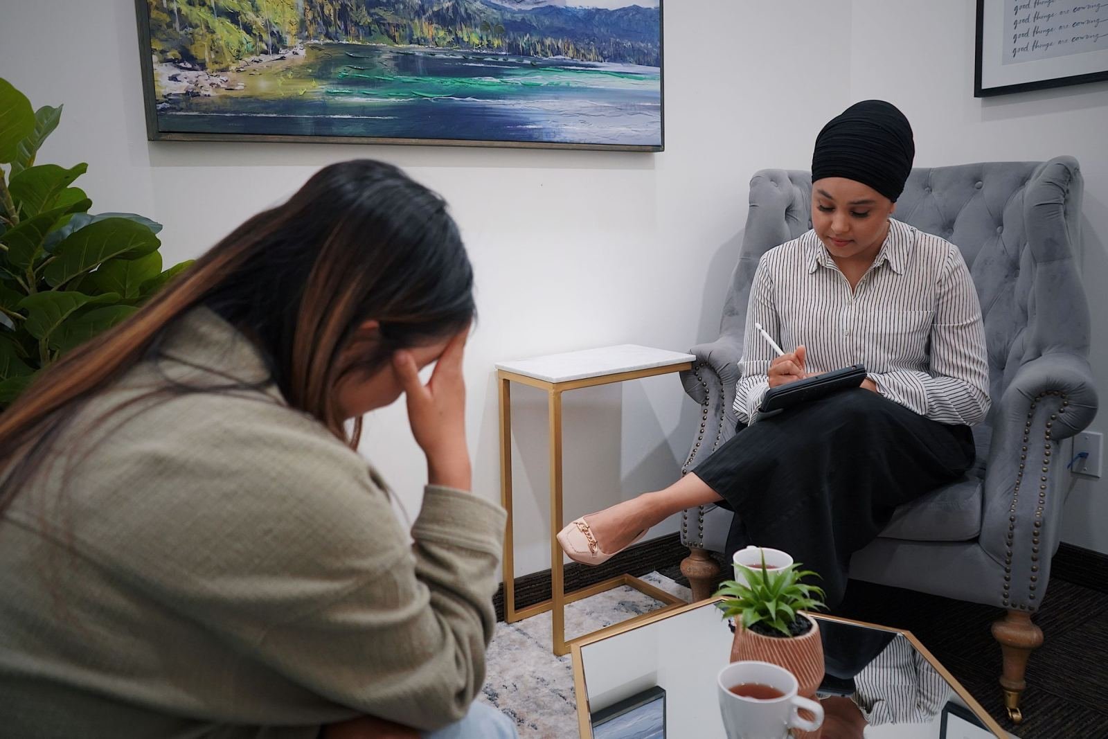A clinician in a grey armchair takes notes while speaking with a client