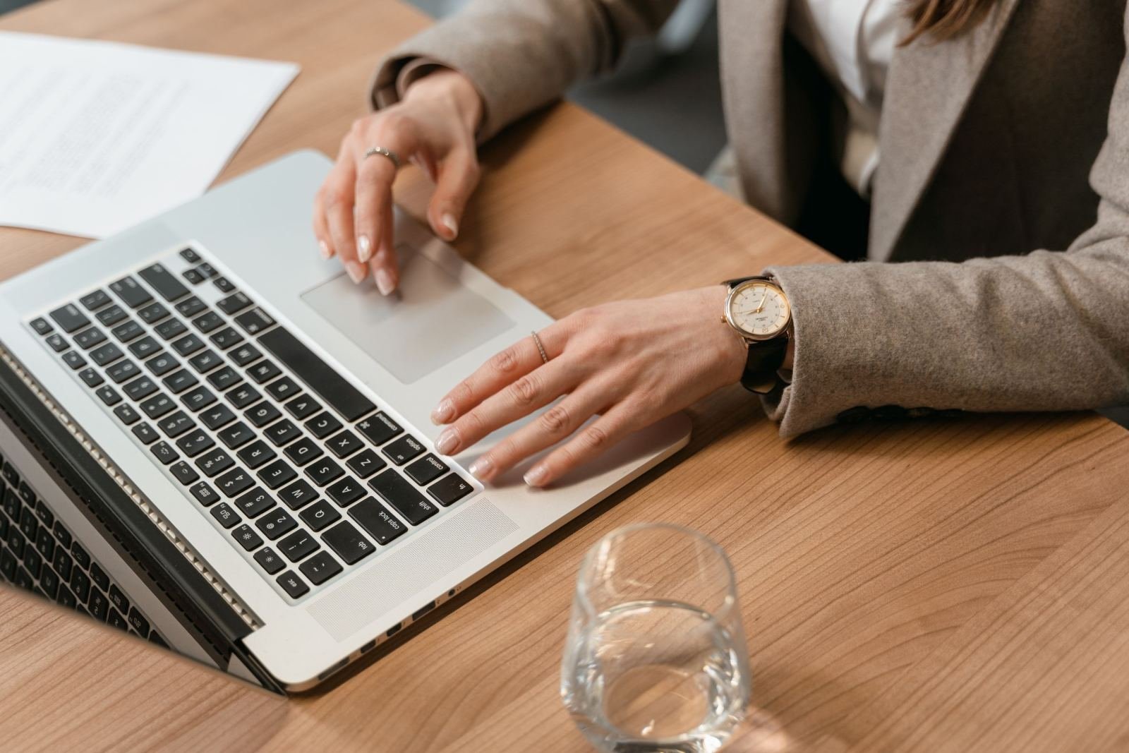 A person in a blazer uses a laptop at a wooden table with a glass of water