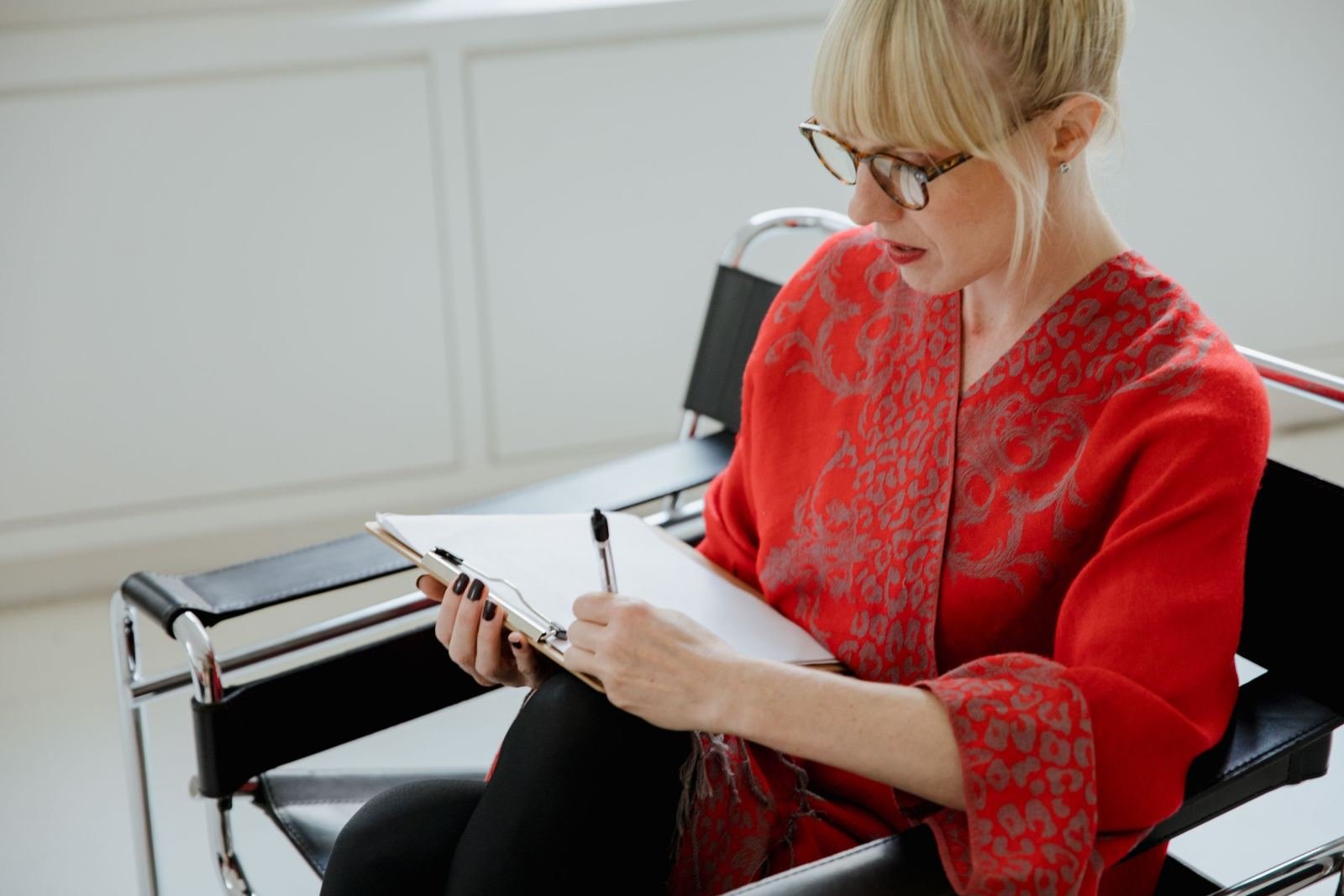 A woman in a red jacket writes on a clipboard while sitting in a black chair