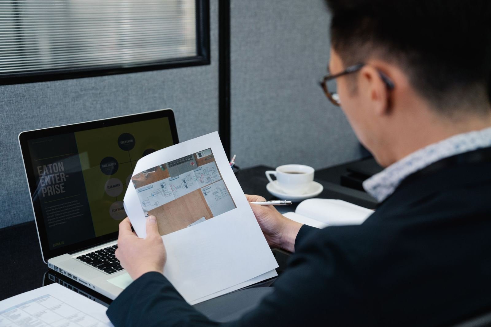A man in a blazer reviews a printed floor plan at a desk with a laptop