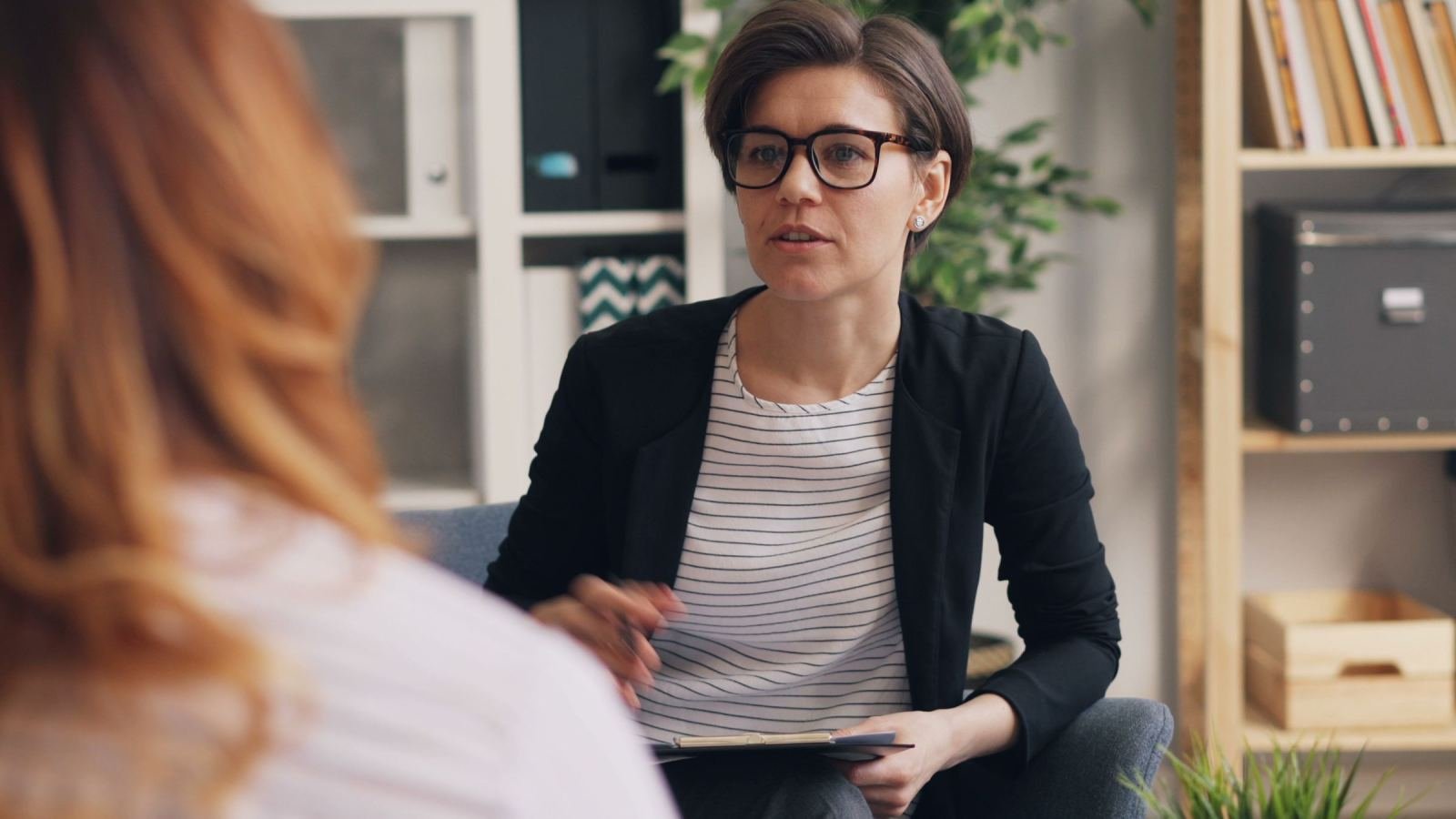 A woman in a black blazer speaks with a person in a bright office