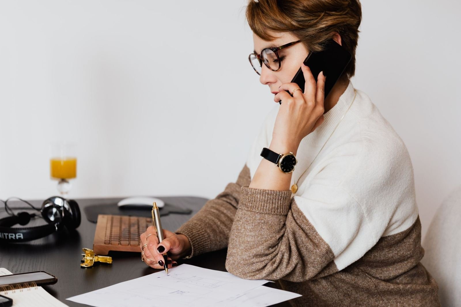 A woman wearing glasses talks on a phone while writing on a document at her desk