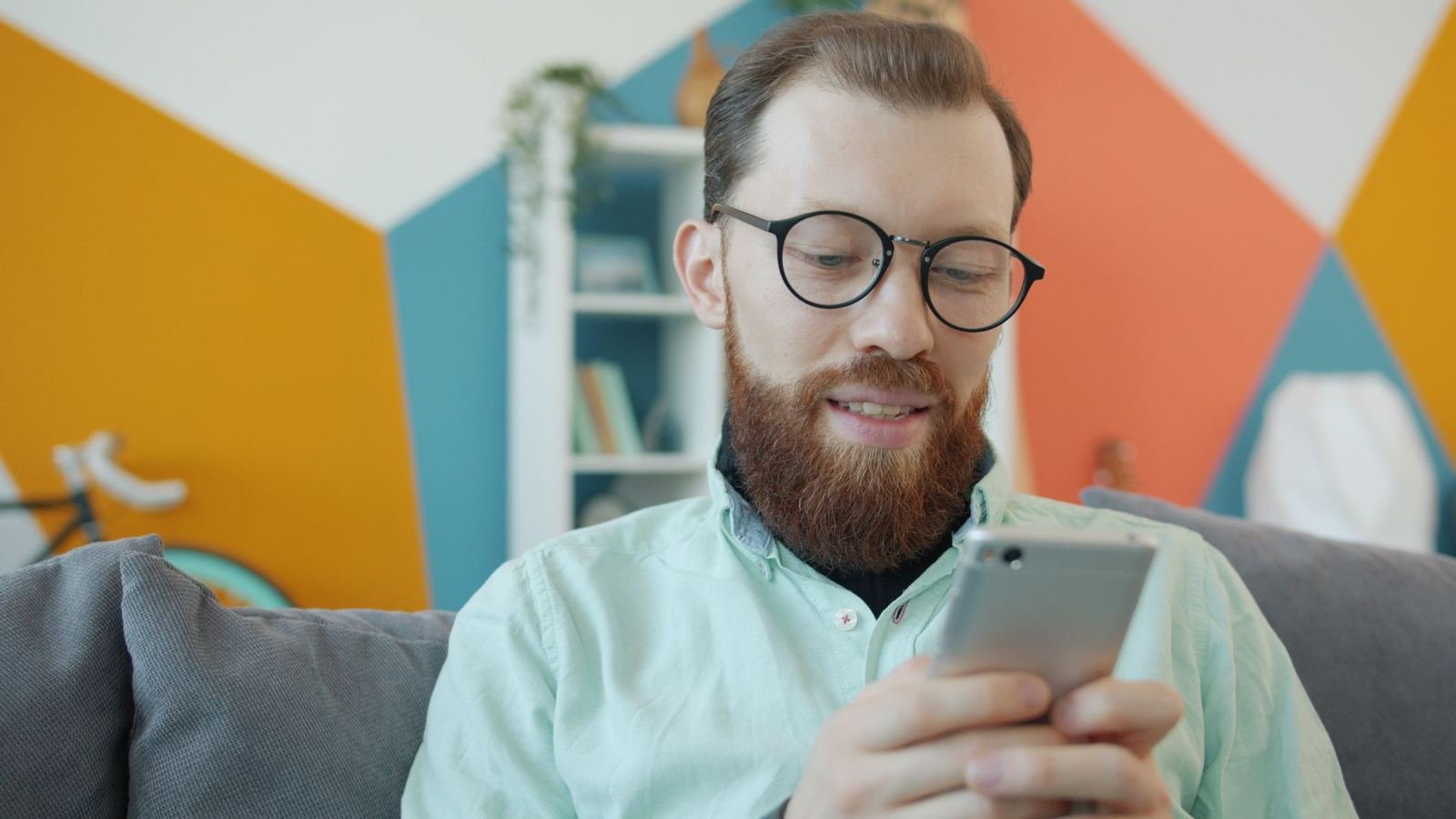 A bearded man with glasses looks at his smartphone while sitting on a sofa