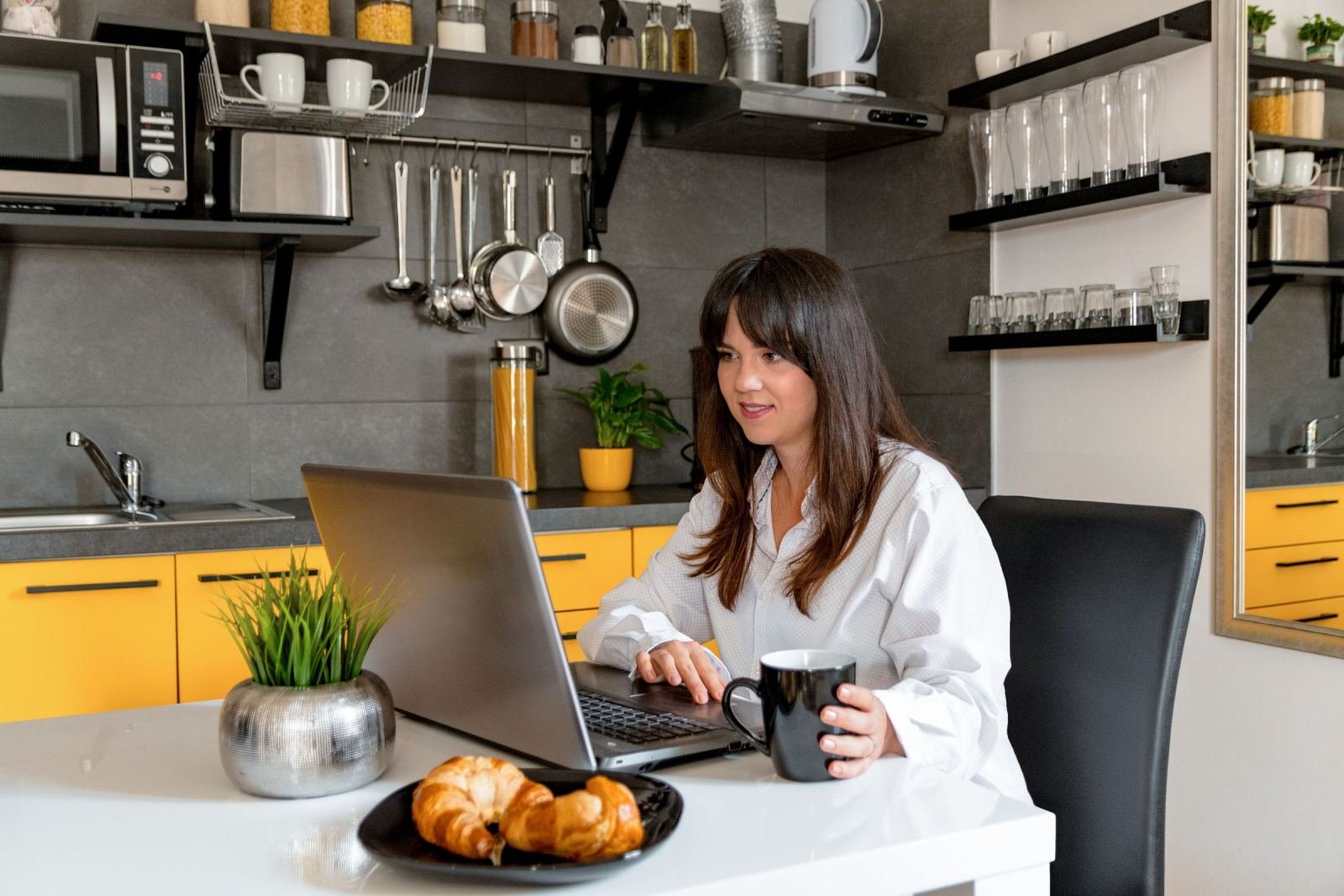 A woman uses a laptop at a kitchen table with a mug and croissants