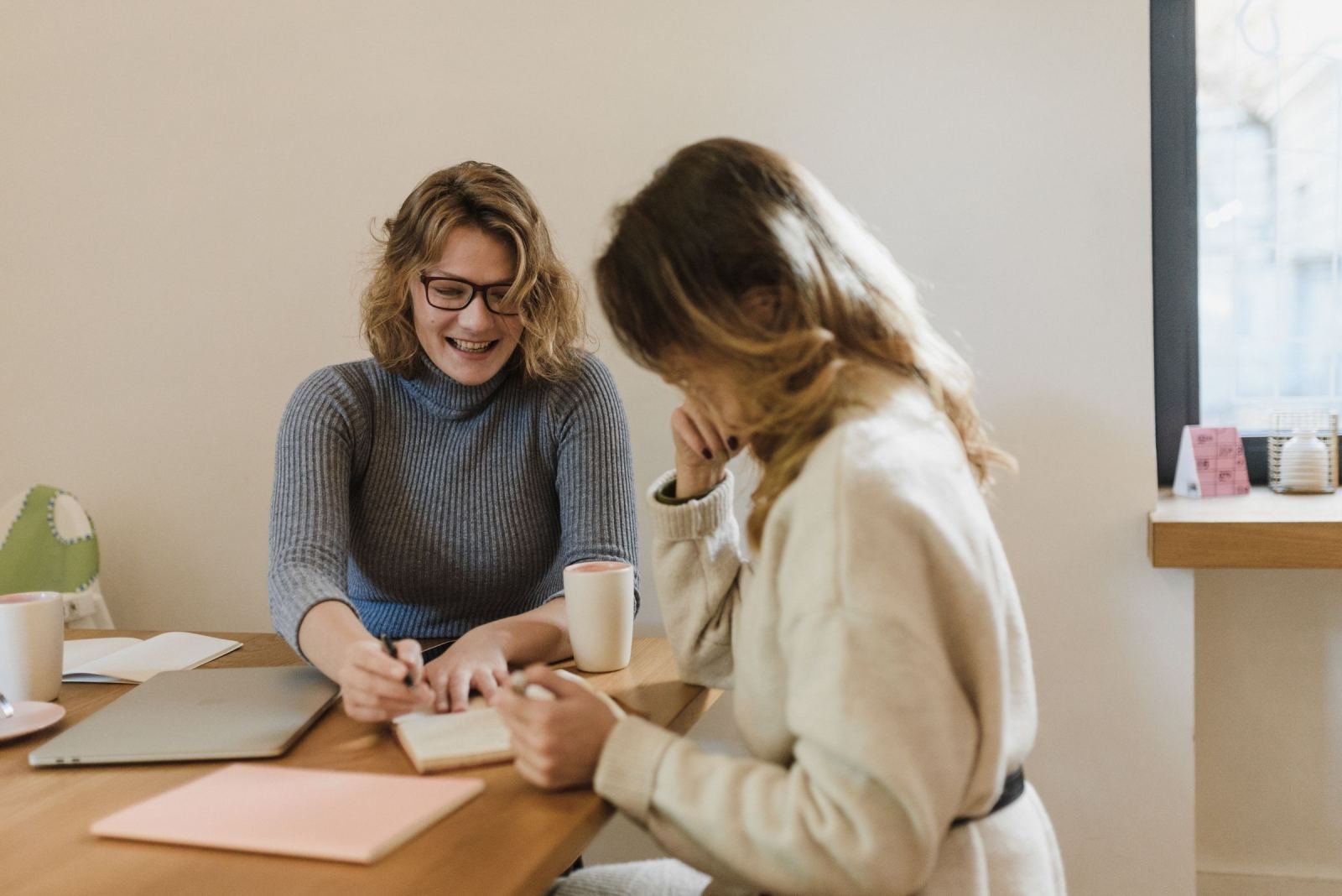 Two women sit at a table together with notebooks and mugs