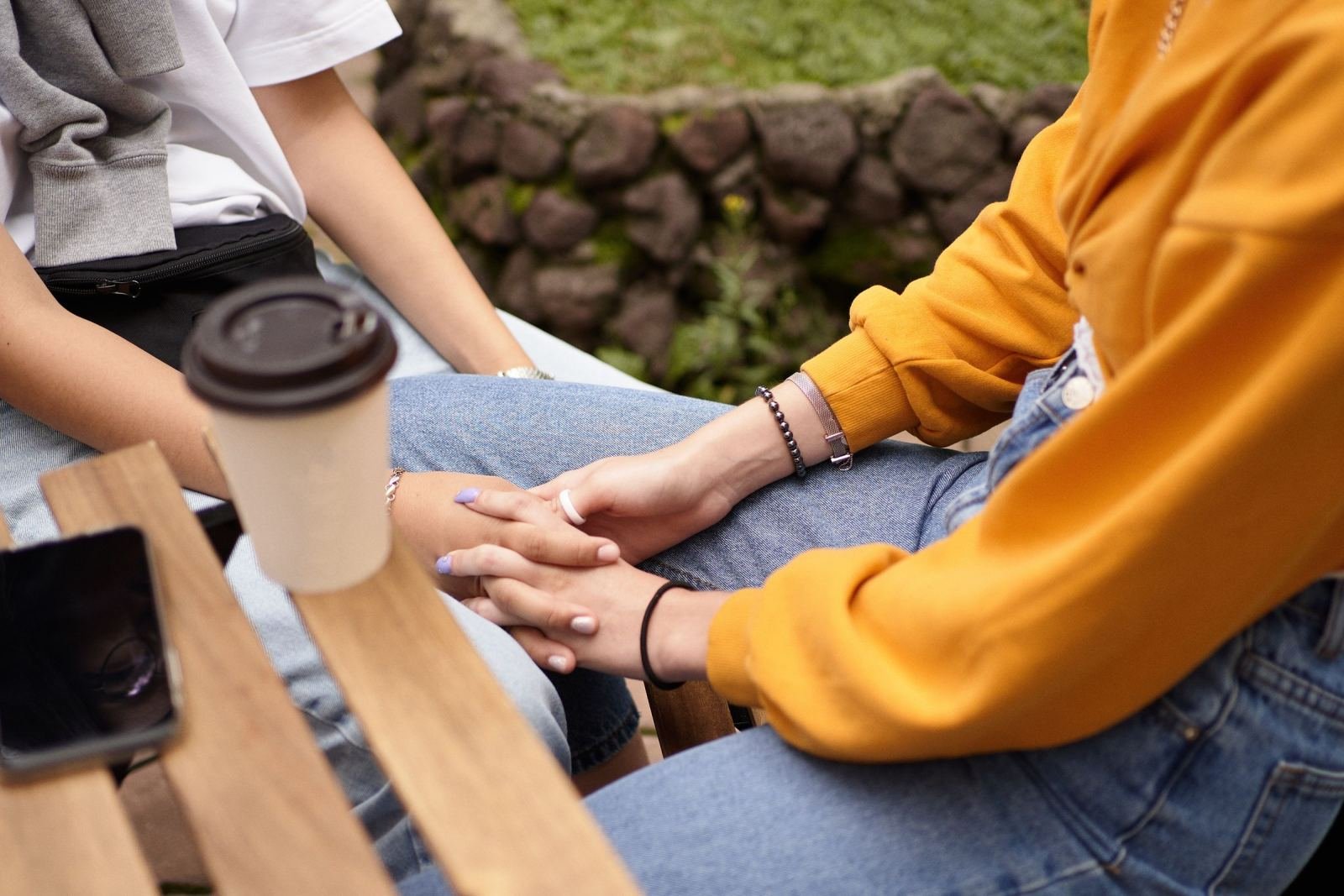 A pair of people sit on a park bench, holding hands. A coffee cup rests nearby.