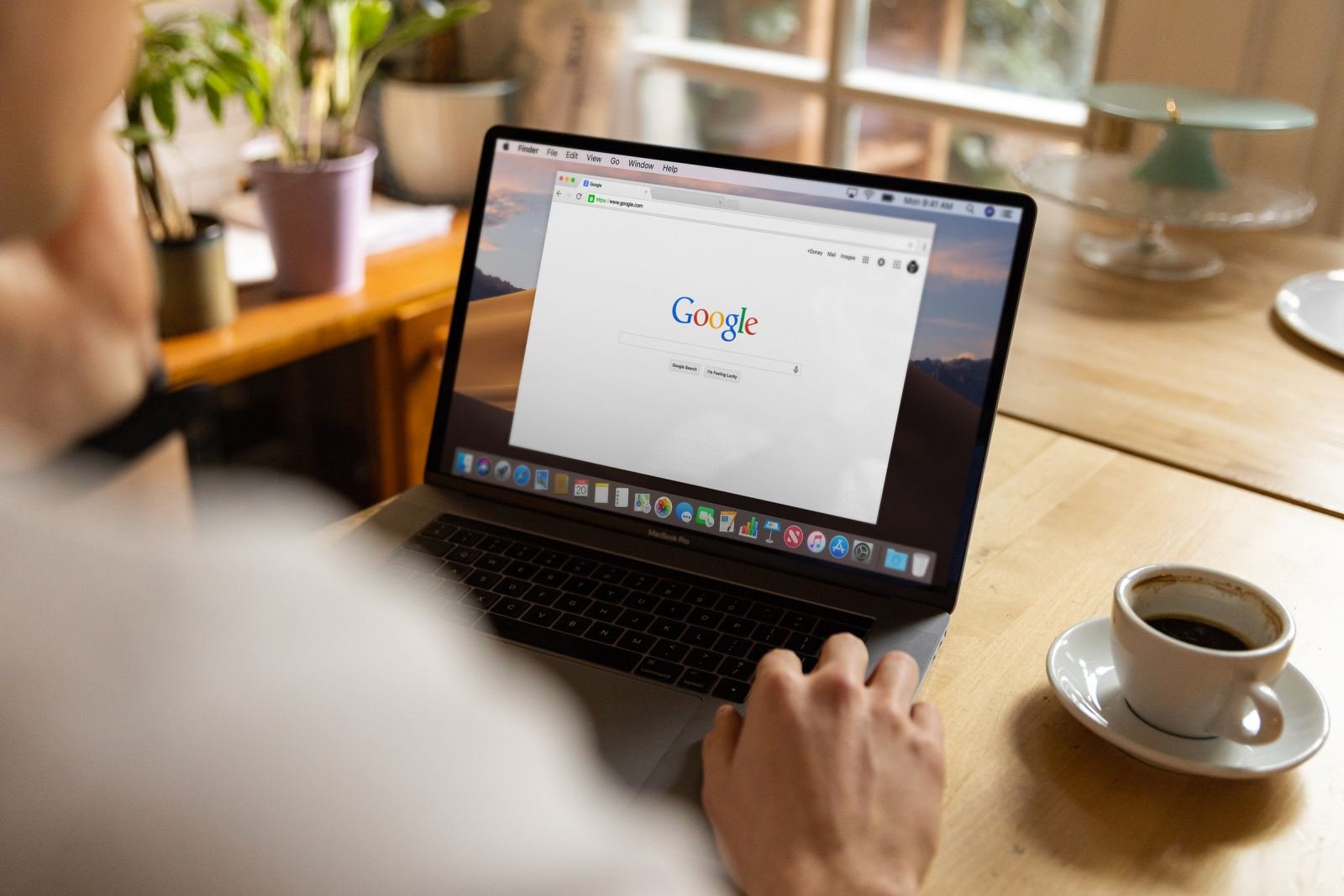 Someone sits at a wooden desk with coffee, using a laptop to look things up online.