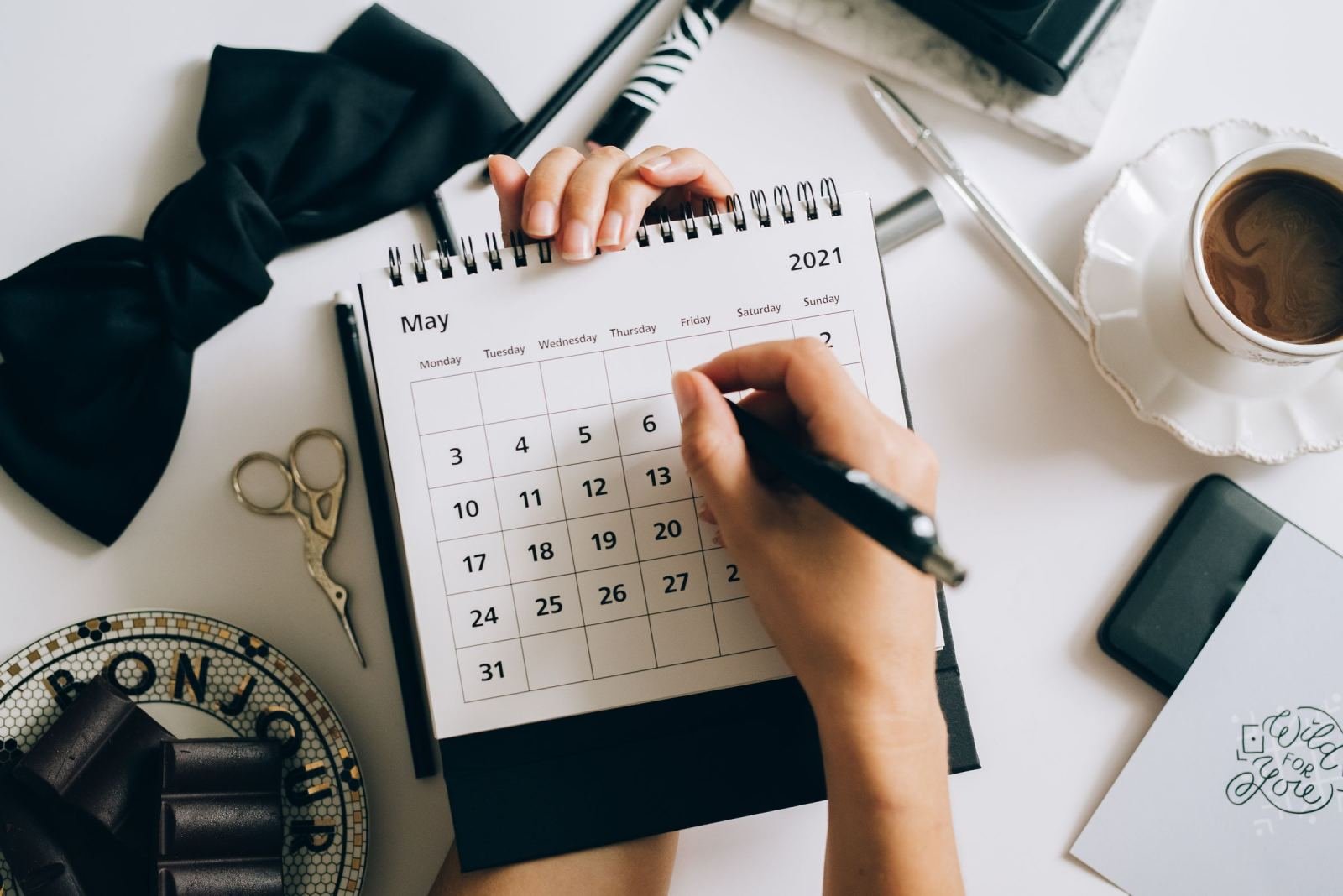 A person writes in a May 2021 calendar on a white desk with coffee and chocolate