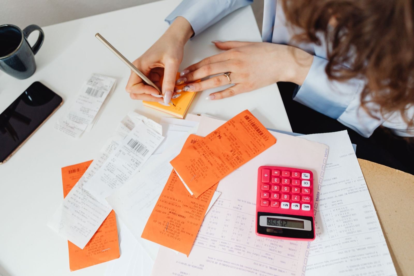 A person writing in a notebook surrounded by receipts and a red calculator