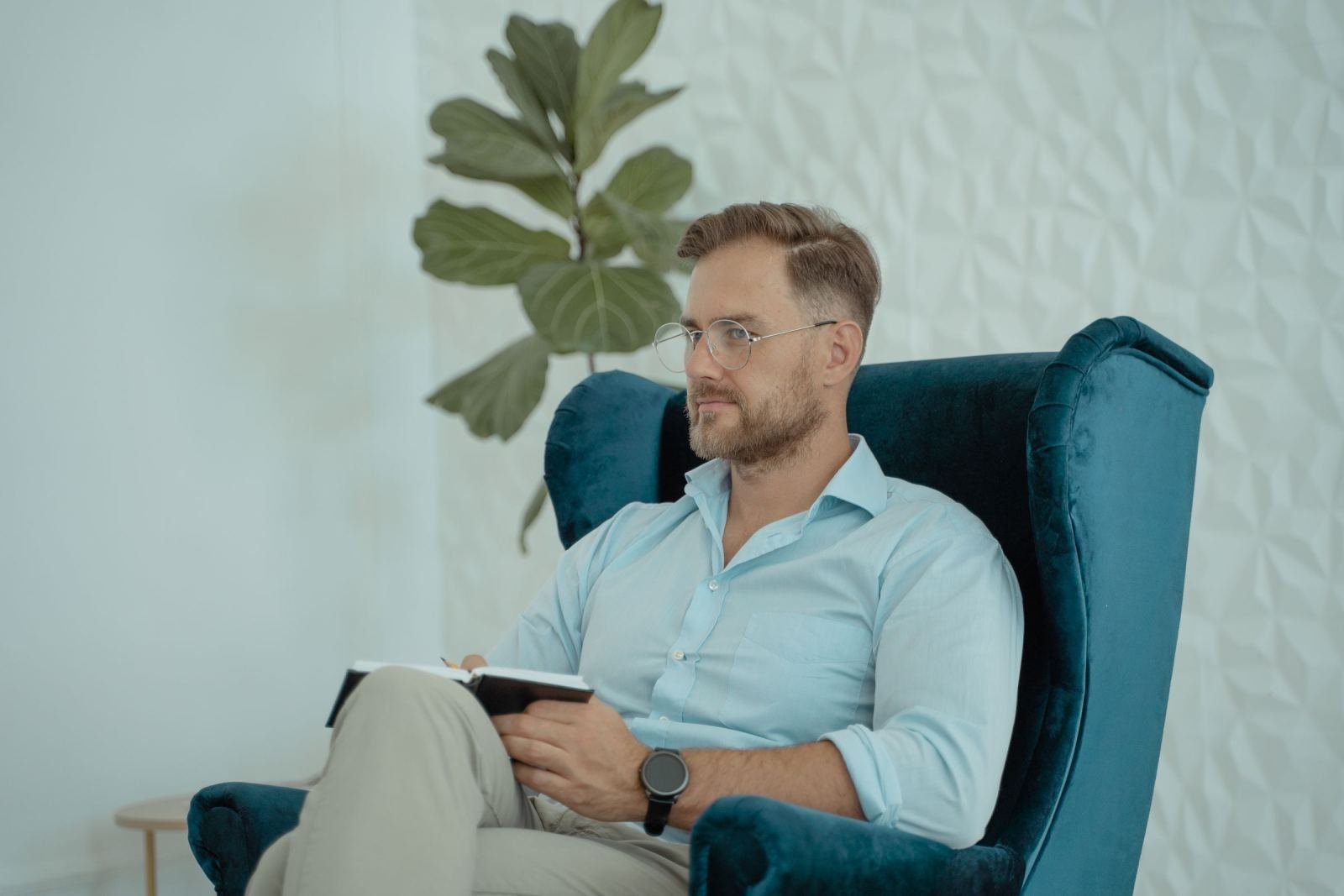 A man with glasses sits in a blue armchair holding a notebook and pen