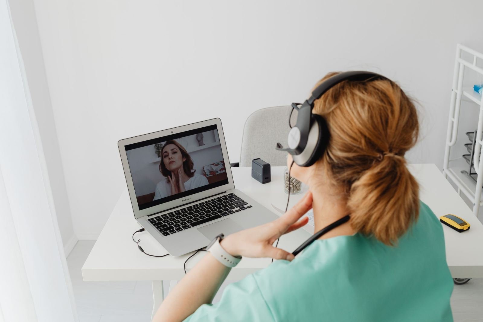 A healthcare worker in scrubs wearing a headset during a video call on a laptop