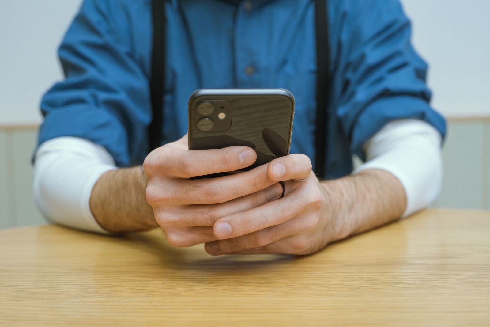 A person in a blue shirt and suspenders holds a smartphone over a wooden table