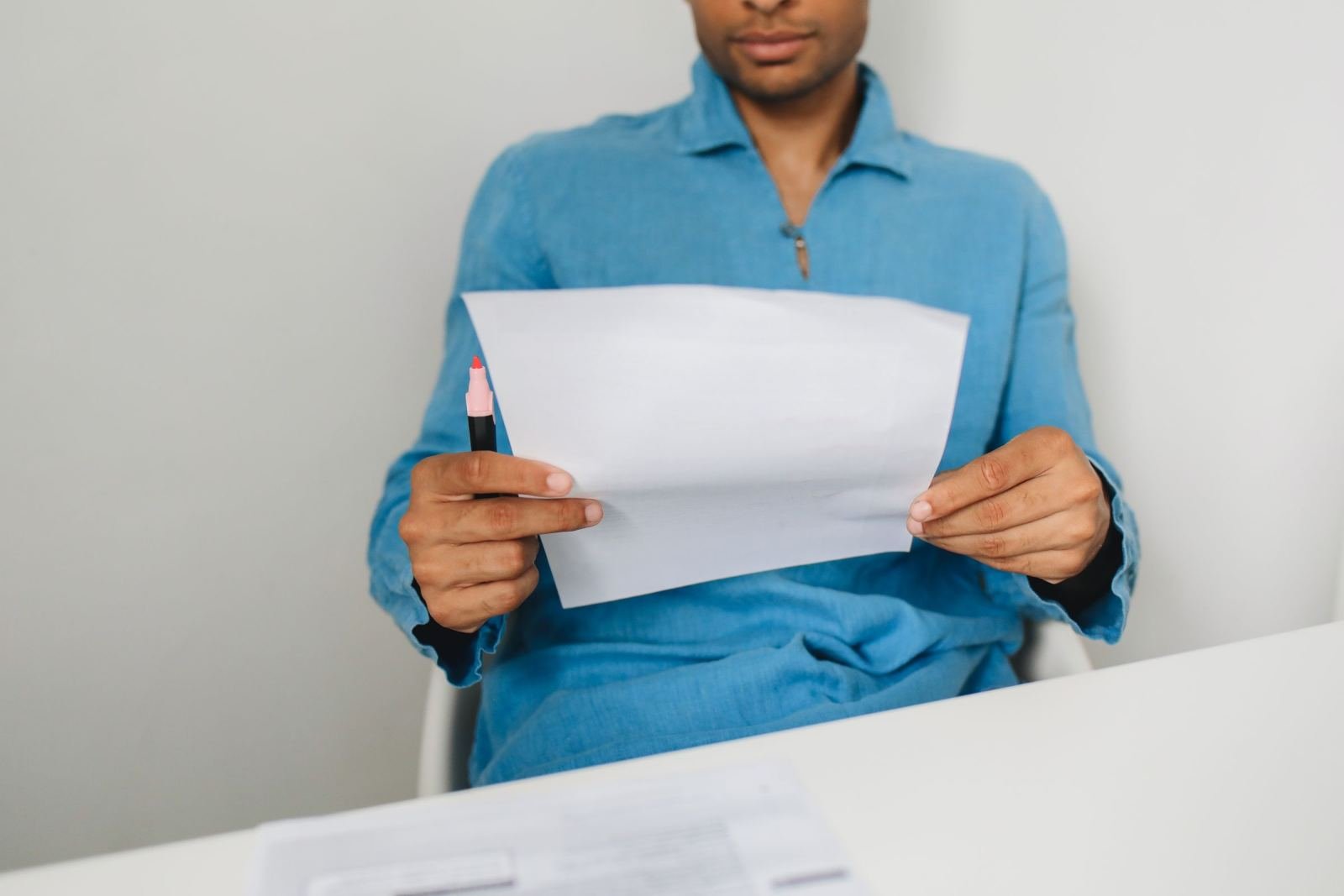 A person in a blue shirt holds a piece of paper and a red pen at a white table