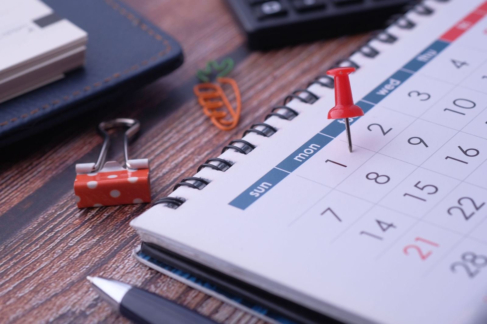 A red pushpin marks a date on a spiral-bound desk calendar