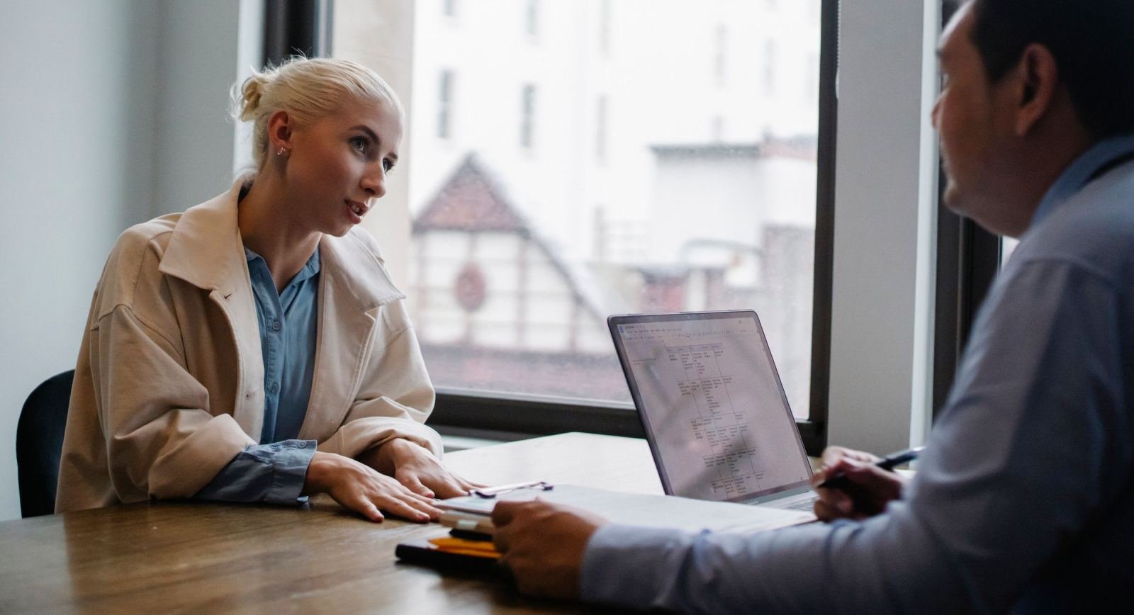A woman in a tan jacket speaks with a man across a table with a laptop