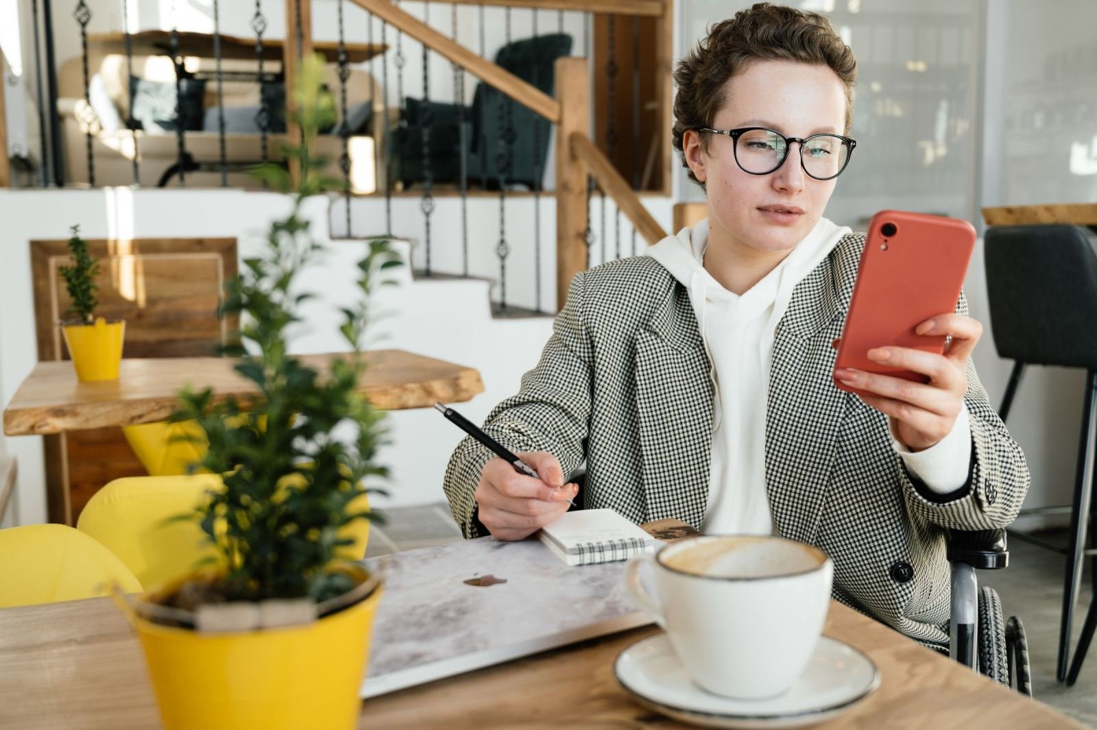 A woman in a blazer looks at her phone while sitting at a cafe table with a notebook and coffee