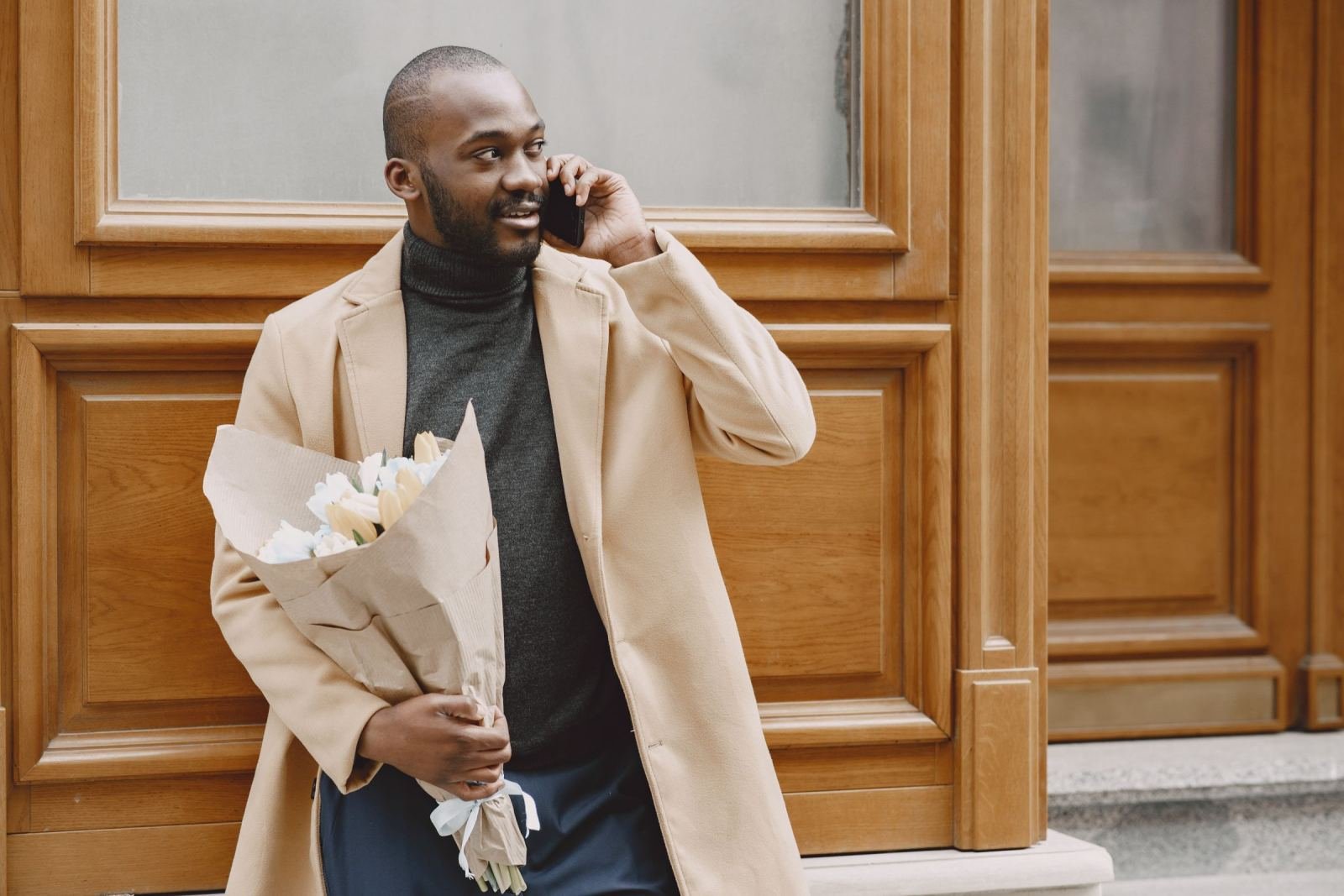 A man in a tan coat talks on a phone while holding a bouquet of white flowers