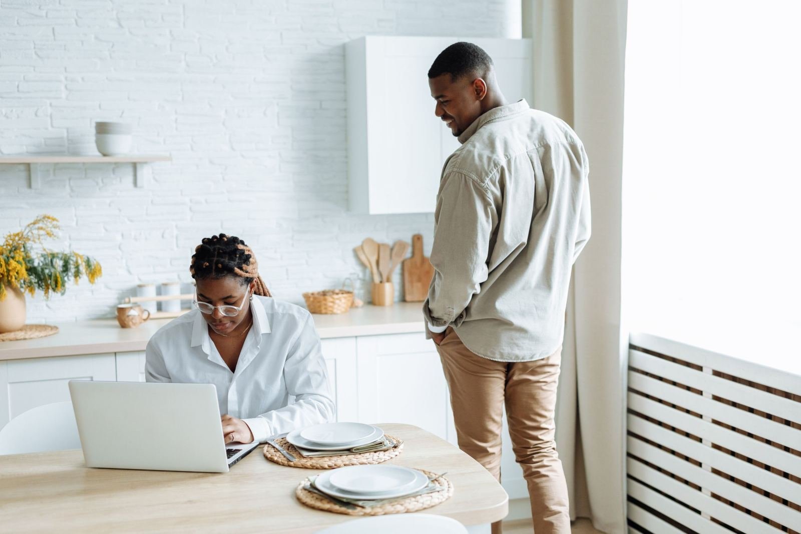 A woman uses a laptop at a kitchen table while a man stands beside her