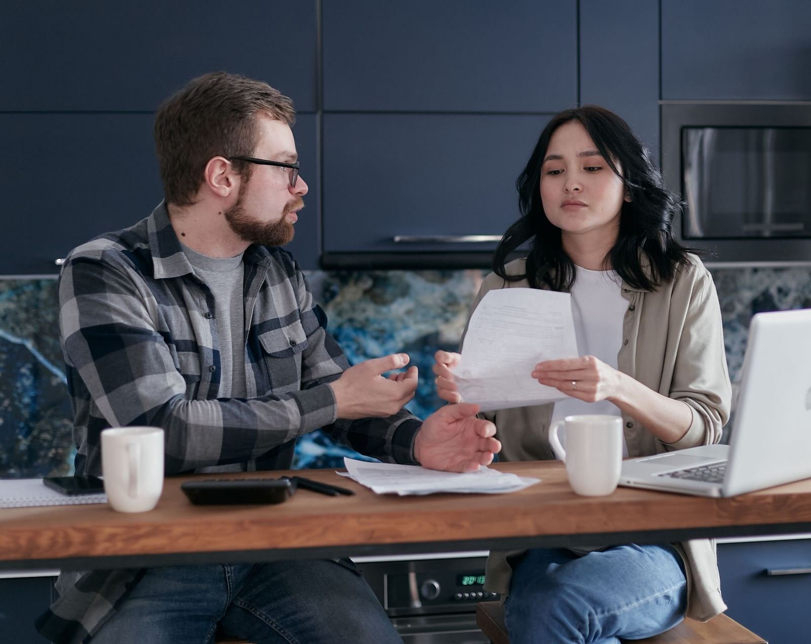 A man and woman review a document together at a kitchen counter with a laptop
