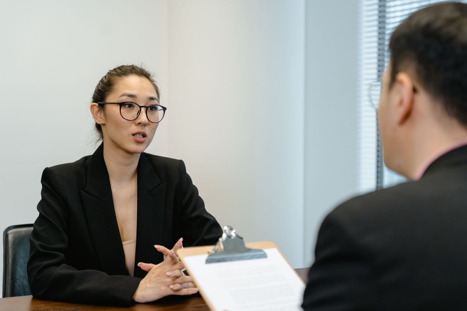 A woman in a black blazer speaks with a man across a table with a clipboard