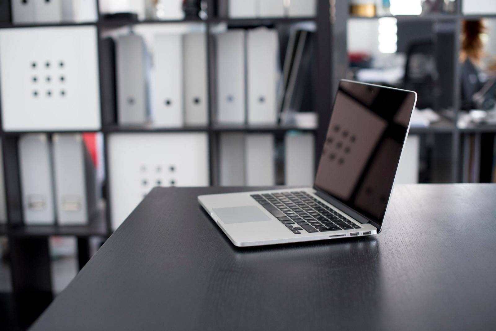 An open laptop on a dark desk with blurred office shelves in the background
