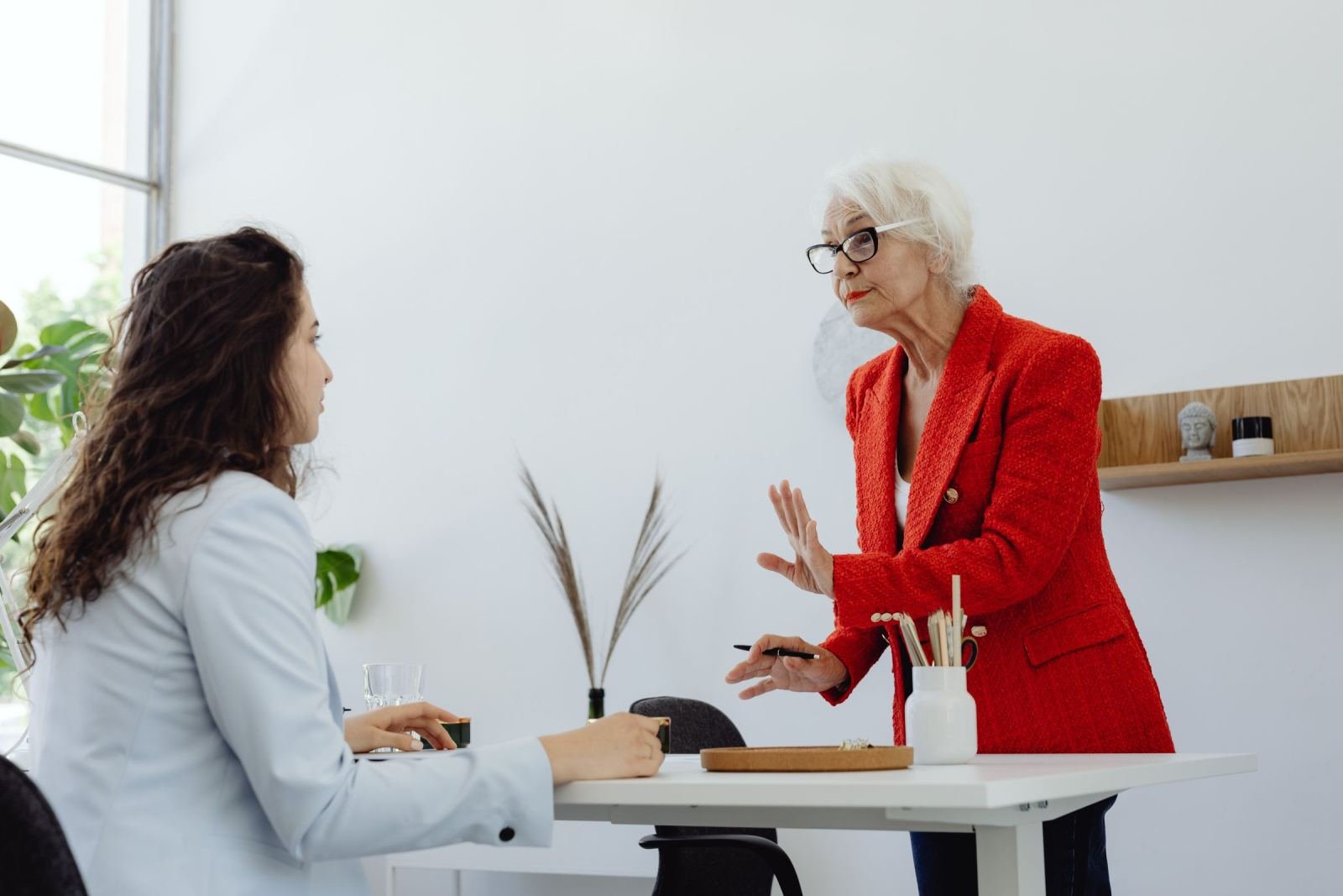 An older woman in a red blazer gestures while talking to a younger woman at a white table