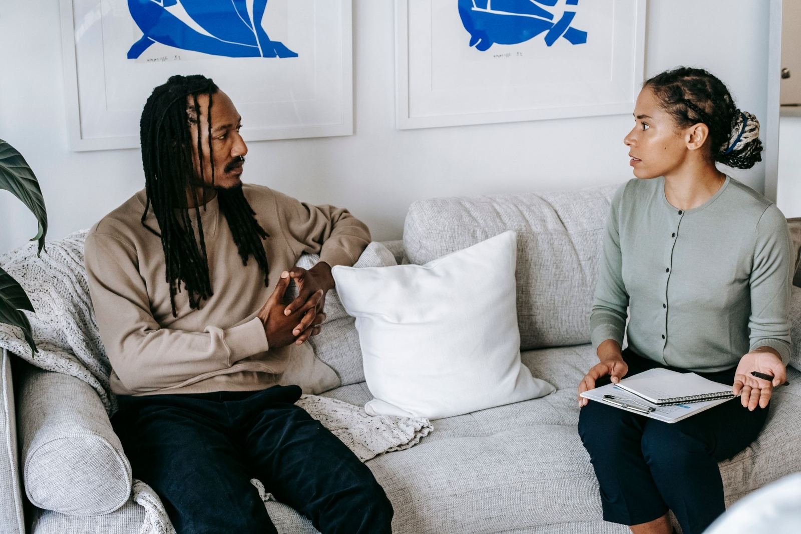 A man and a woman with a notebook talk while sitting on a gray sofa