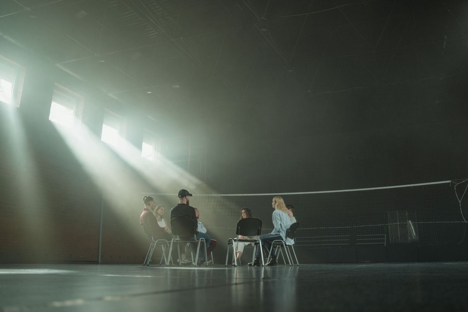 A small group of people sit in a circle of chairs in a dim room with beams of light