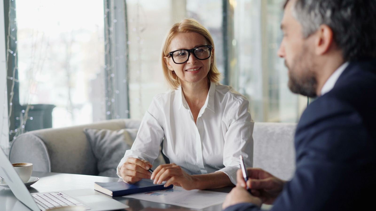A woman wearing a white shirt and glasses is having a conversation with a man in a suit at a table.