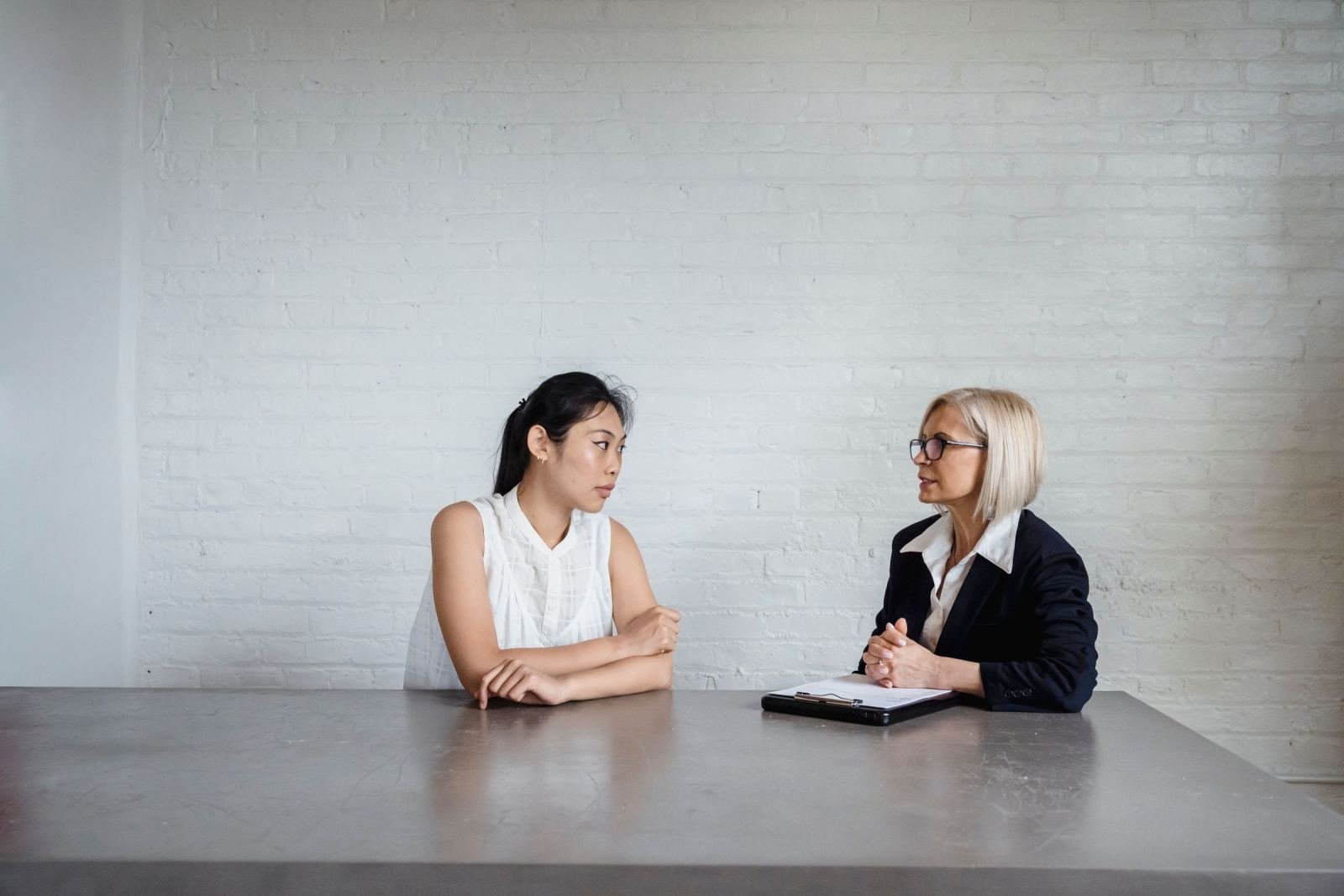 Two women sit at a table talking, with one holding a clipboard