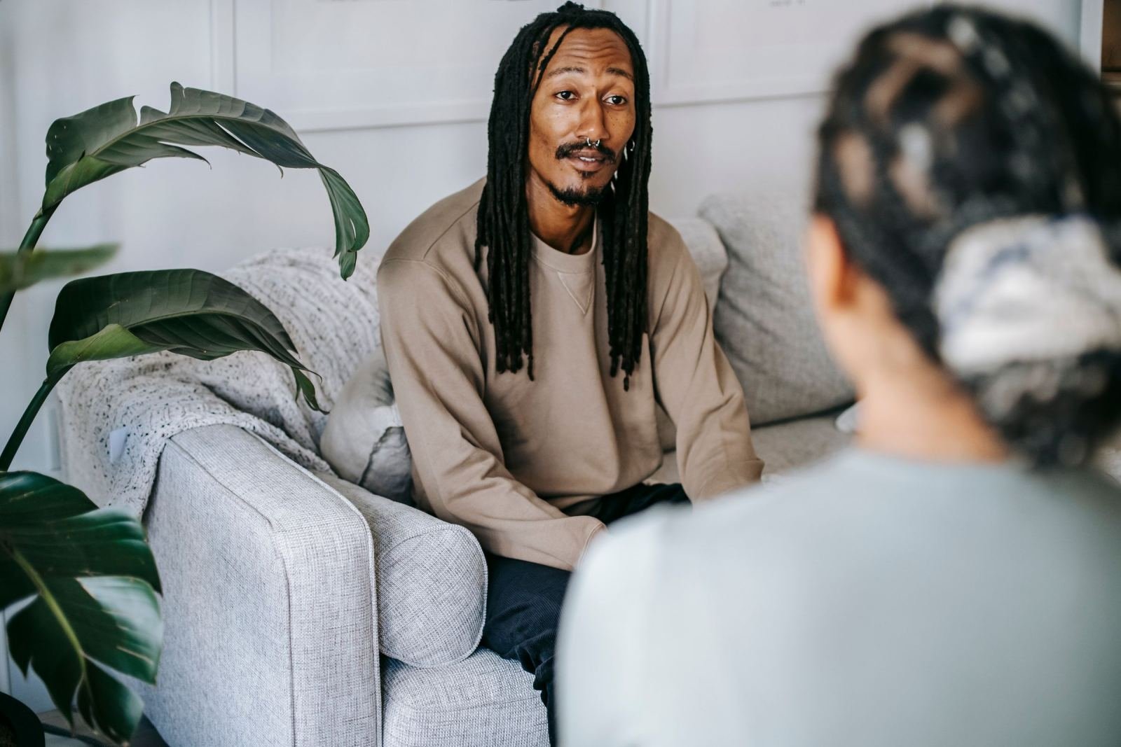 A man with dreadlocks sits on a gray sofa talking to another person