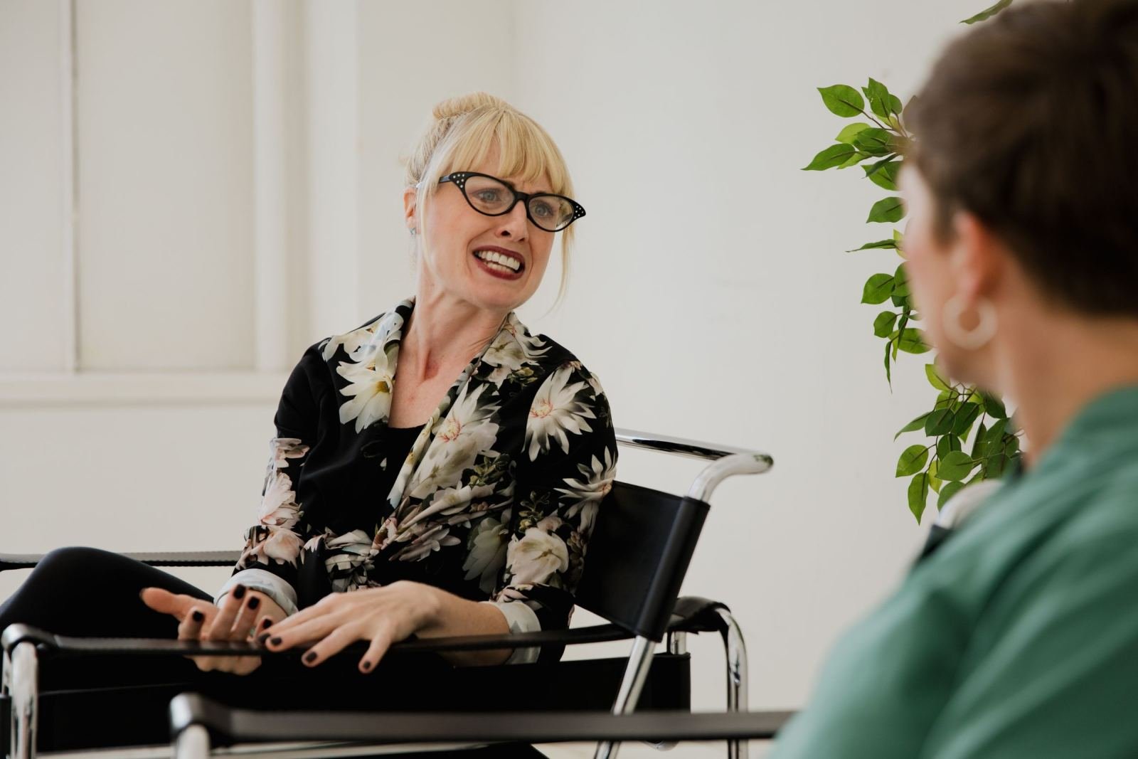 A woman with glasses gestures while talking to another person in a bright office