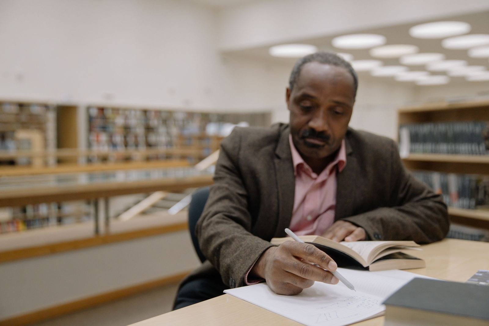 A man in a blazer reads a book and writes in a notebook at a library table