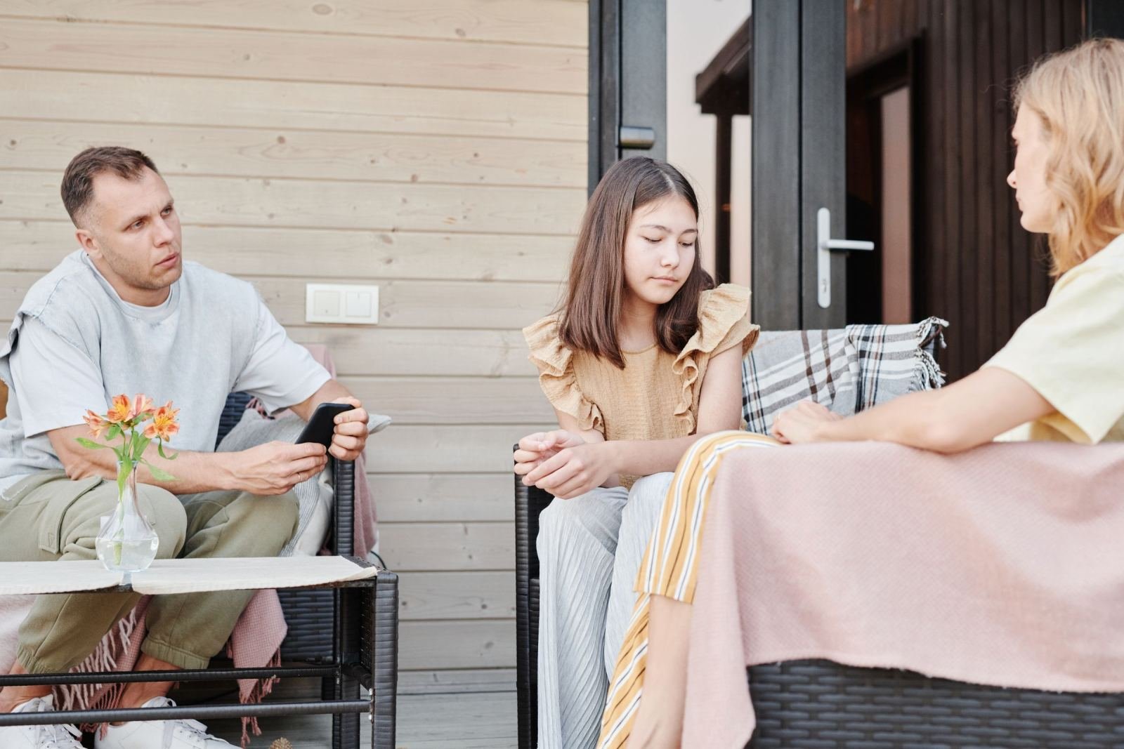 A man, a teenage girl, and a woman sit together on a wooden deck