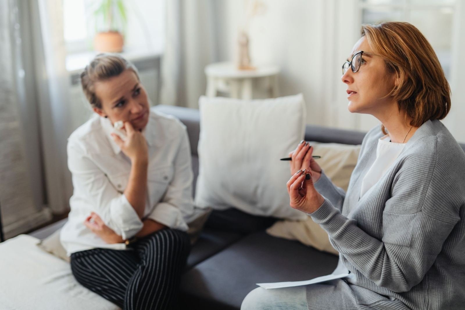 A woman in glasses speaks with a pen in her hand to another woman on a sofa