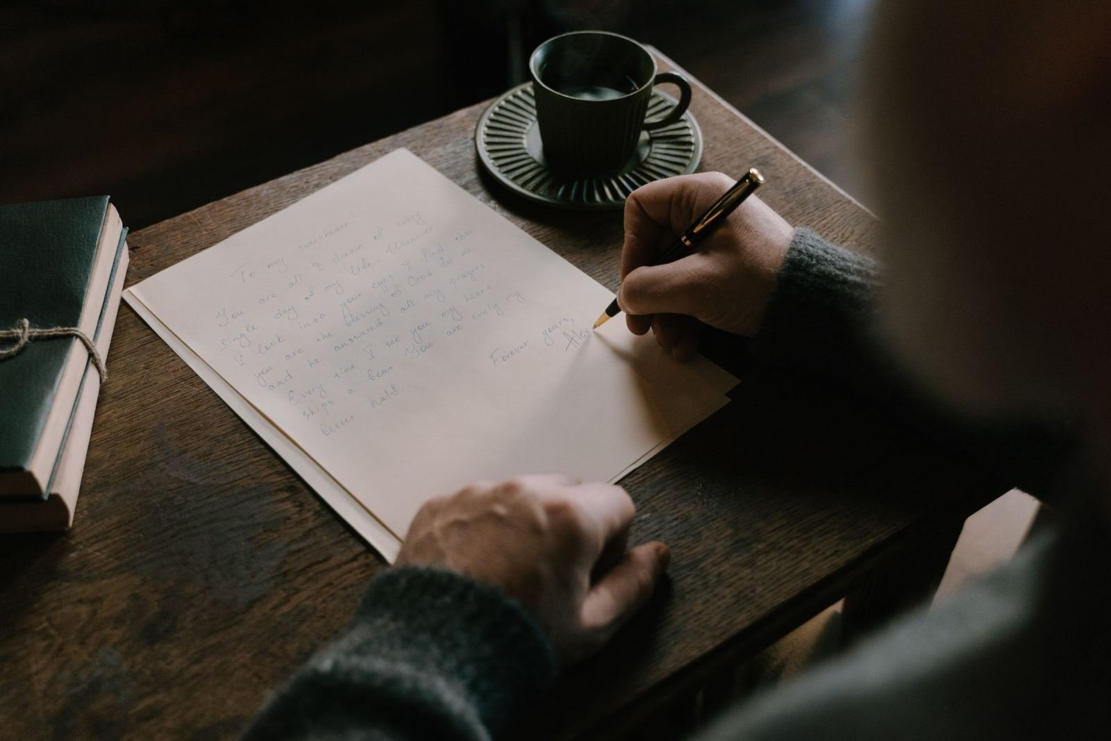 A person writing a handwritten letter on a wooden table next to a mug