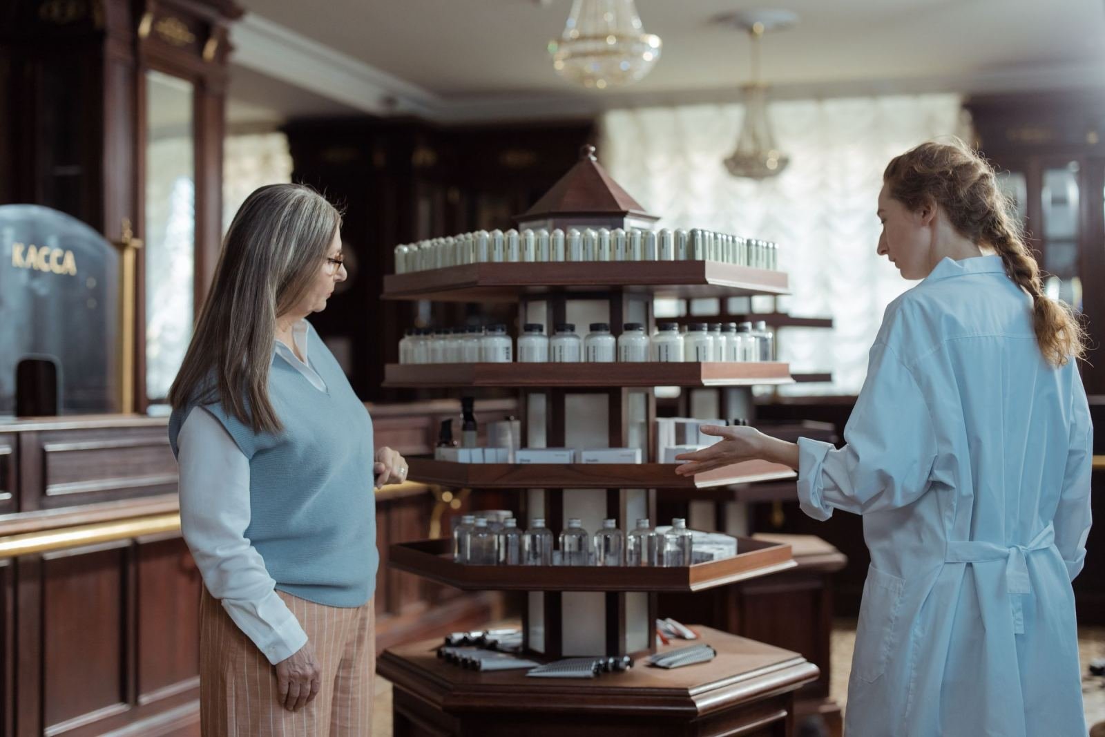 A pharmacist wearing a white coat chats with a client beside a row of medicine bottles.
